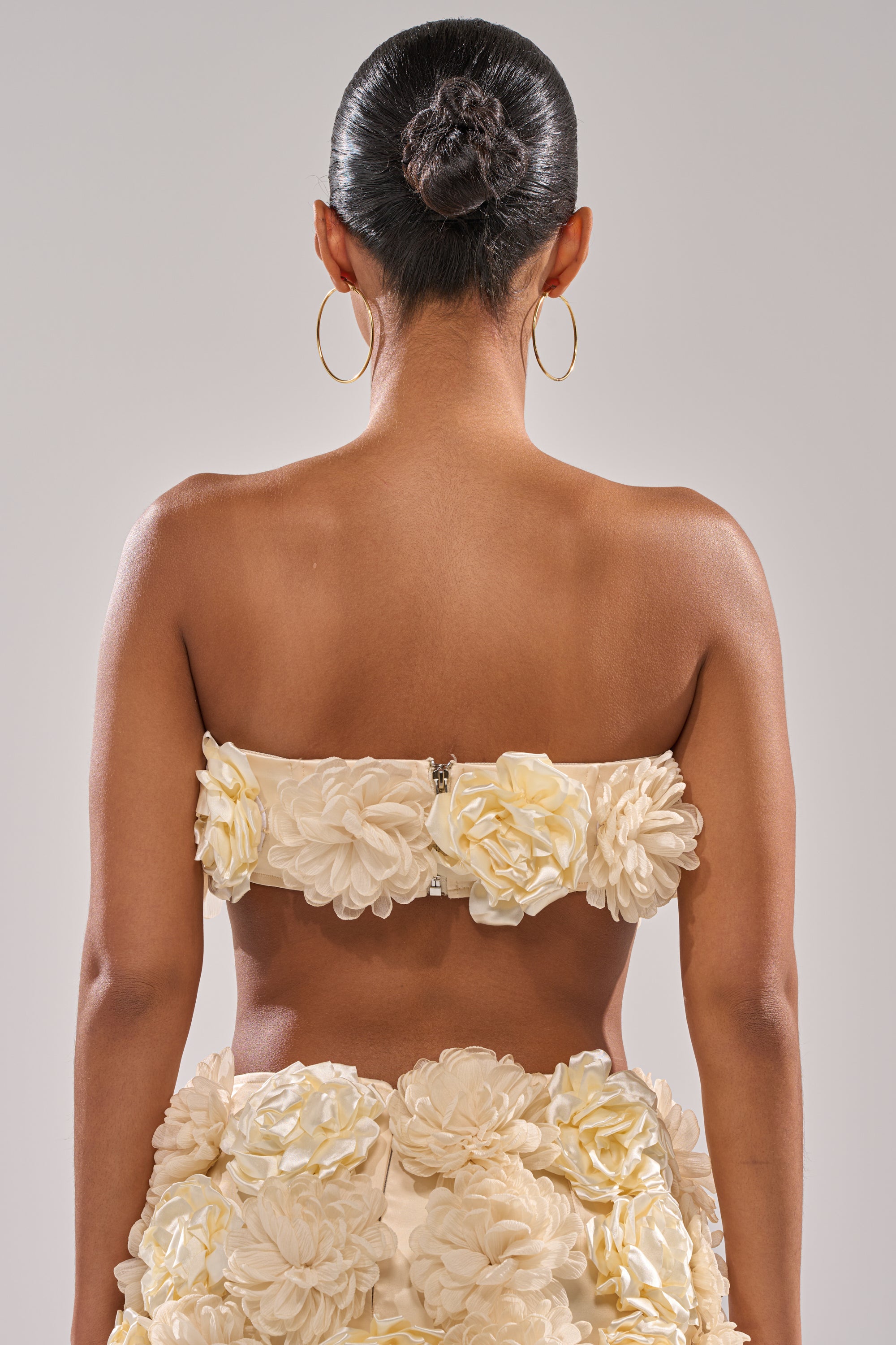 A woman with her hair in a sleek bun is seen from behind, wearing large hoop earrings and the FLOWER POWER BRALETTE—a cream satin strapless top decorated with striking 3D floral appliqués. 8
