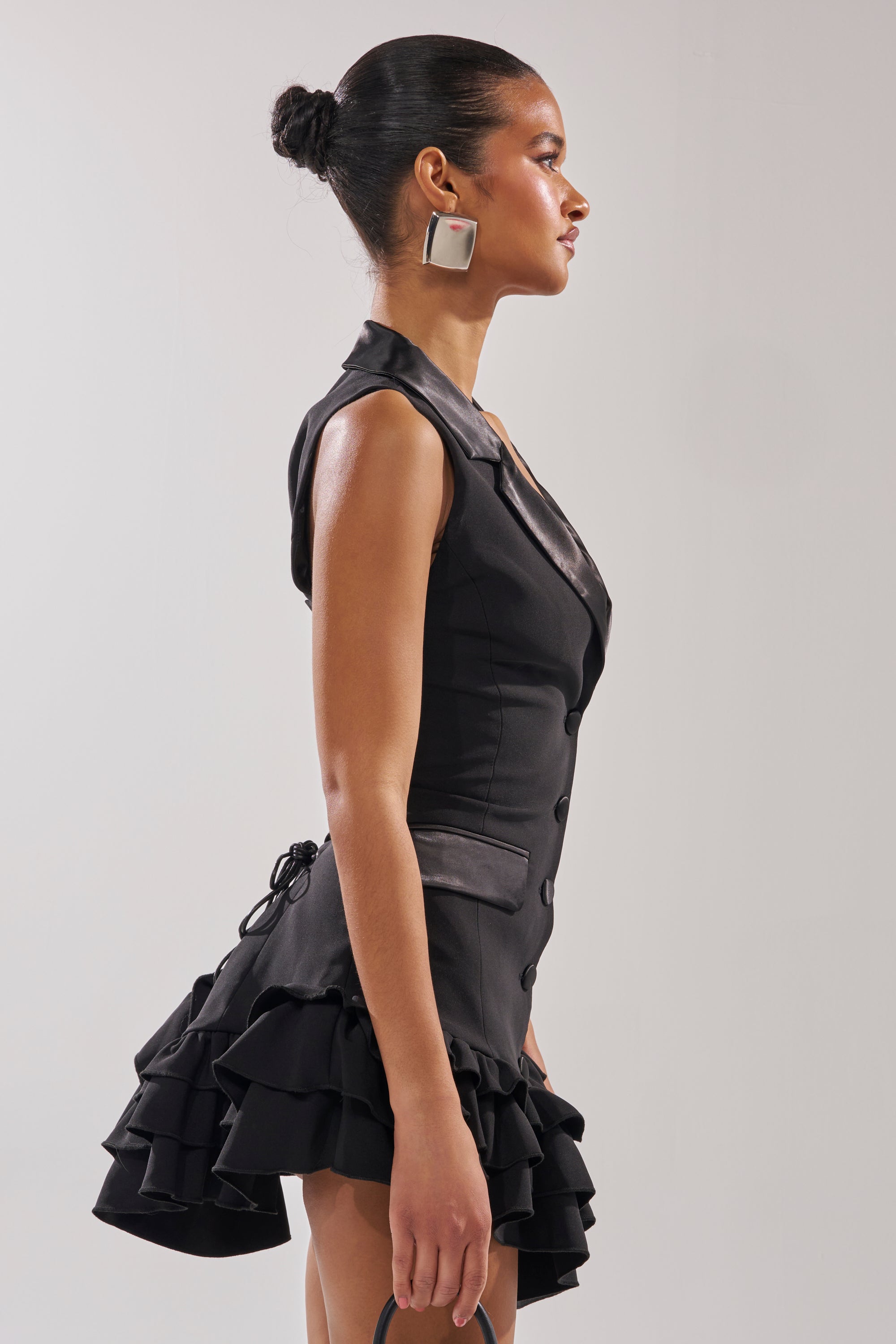A woman stands in profile wearing the SUITED UP SLEEVELESS MINI DRESS, a blazer-inspired sleeveless mini with a ruffled hemline, paired with large square earrings and her hair styled in a sleek bun.