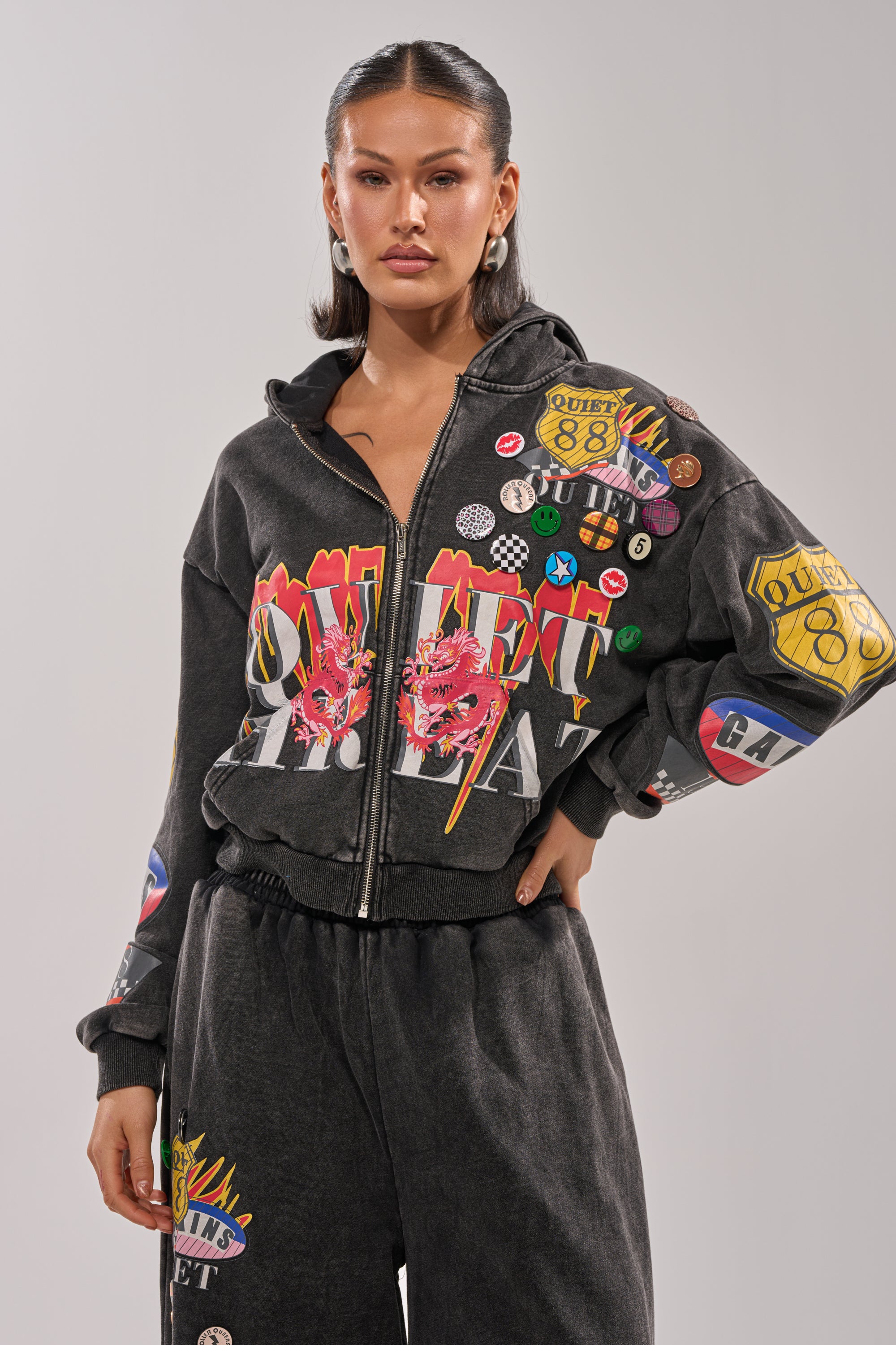 A woman stands confidently with one hand on her hip, wearing the ANOTHER ONE ZIP UP SWEATSHIRT—a black graphic hoodie with colorful prints and pins—paired with matching pants, set against a plain background. 0