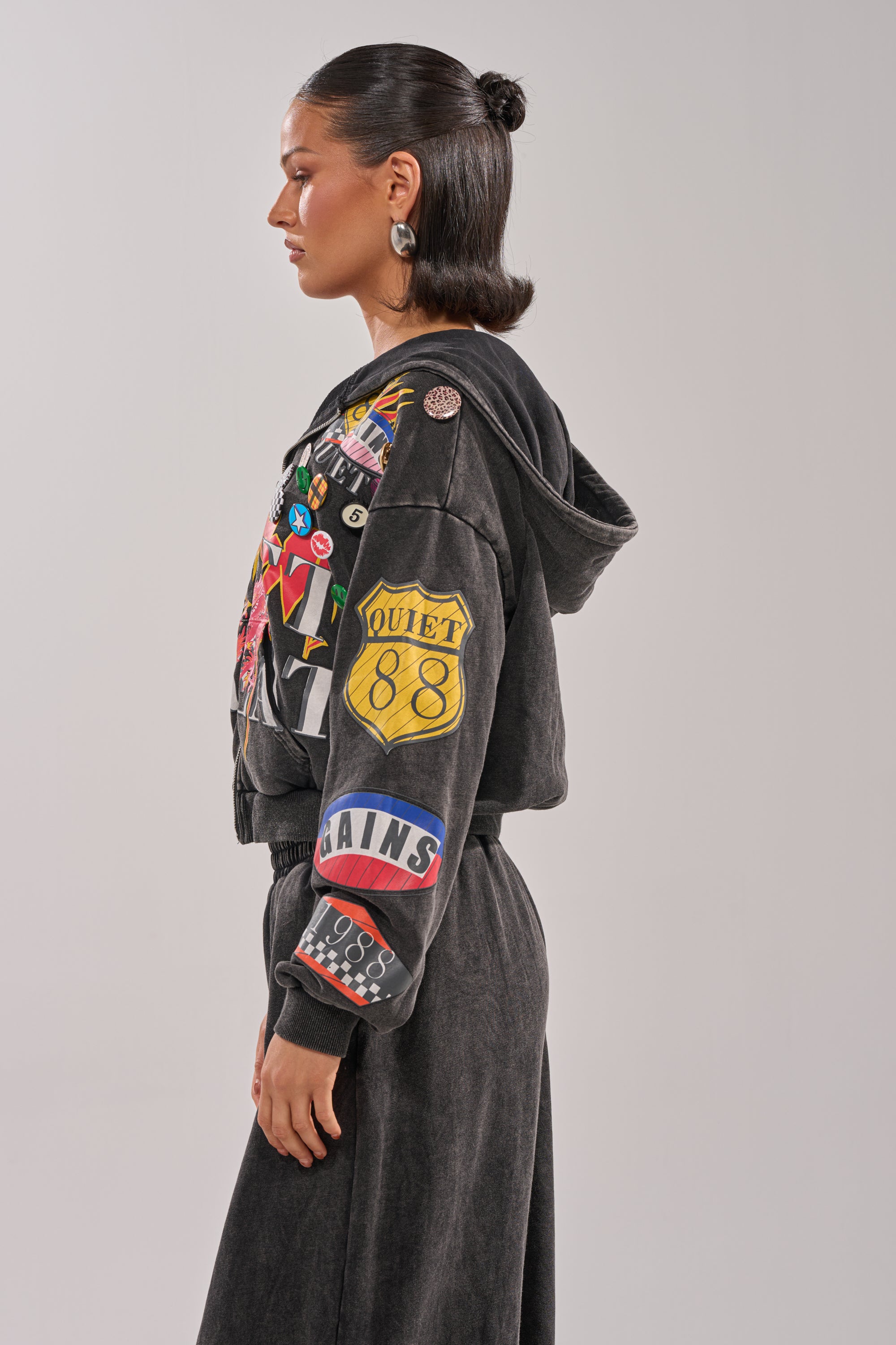 A woman with slicked-back dark hair stands in profile against a plain light background, wearing the ANOTHER ONE ZIP UP SWEATSHIRT decorated with colorful patches and paired with matching black pants. 1