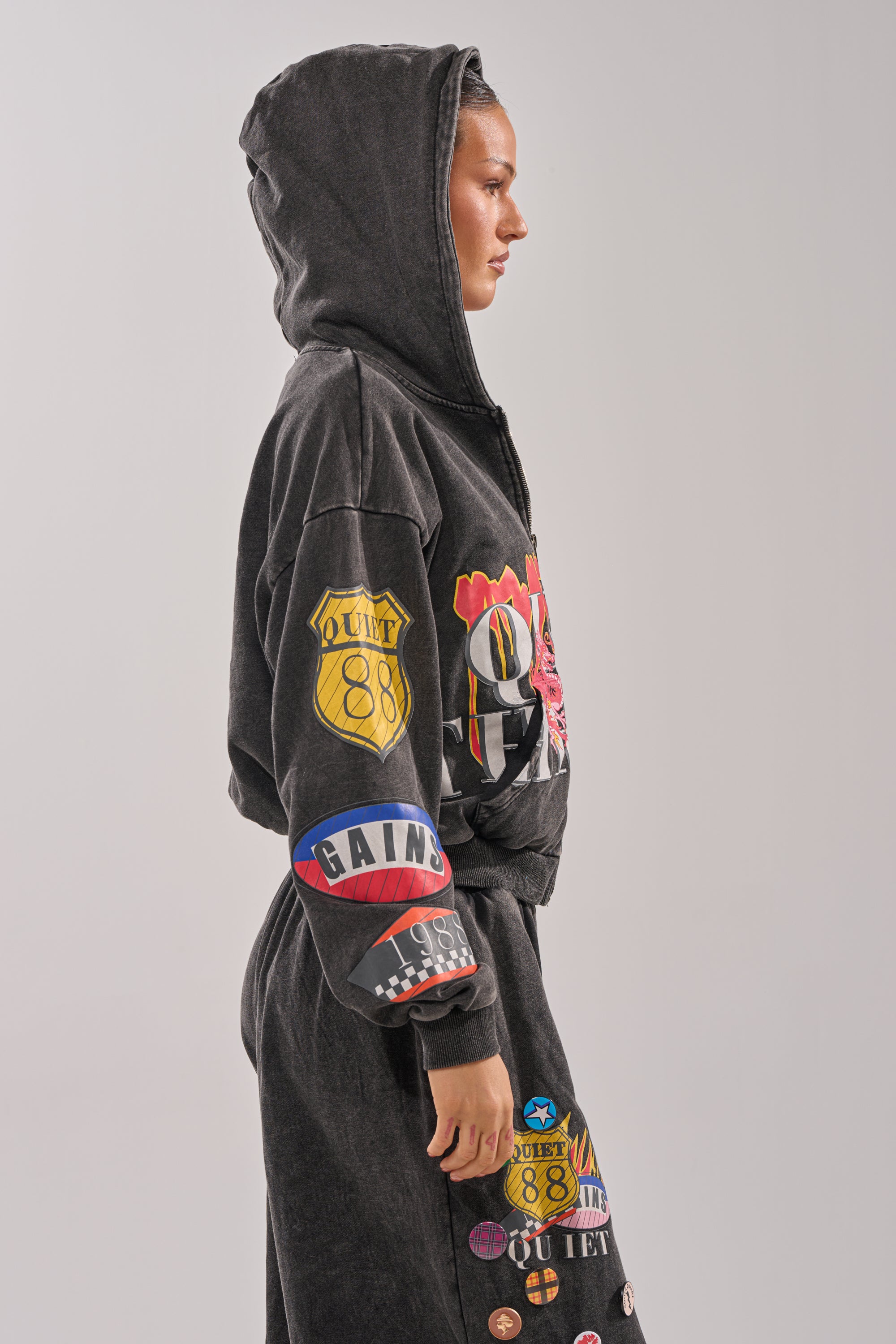A person wearing the ANOTHER ONE ZIP UP SWEATSHIRT and matching pants with bold graphic patches and badges stands in profile against a plain light background. 2