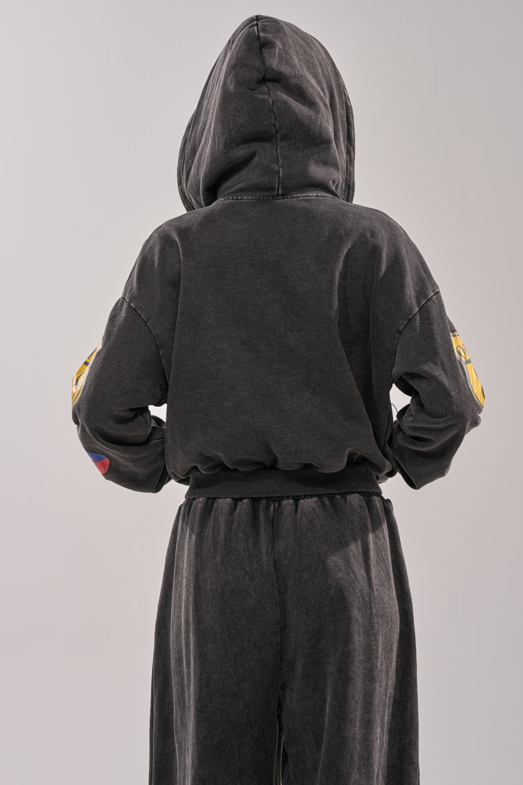 A person stands with their back to the camera, wearing the ANOTHER ONE ZIP UP SWEATSHIRT in black with matching pants. The hood is up, covering their head, and their hands are placed on their hips. 3