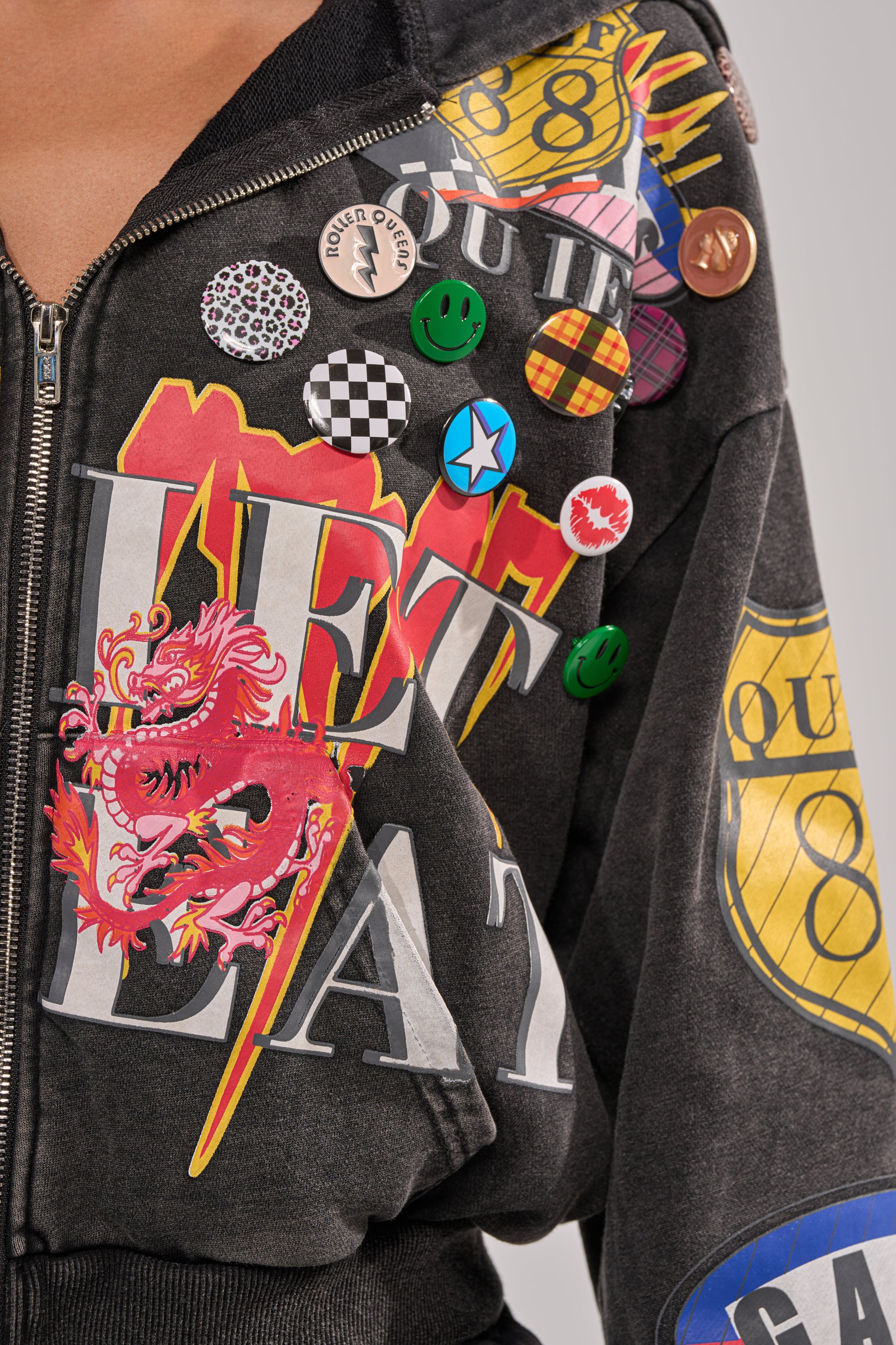 A close-up of the ANOTHER ONE ZIP UP SWEATSHIRT, a black hoodie with colorful designs—featuring a red dragon, yellow shield, and multiple decorative pins—a bold statement piece for streetwear style. 4