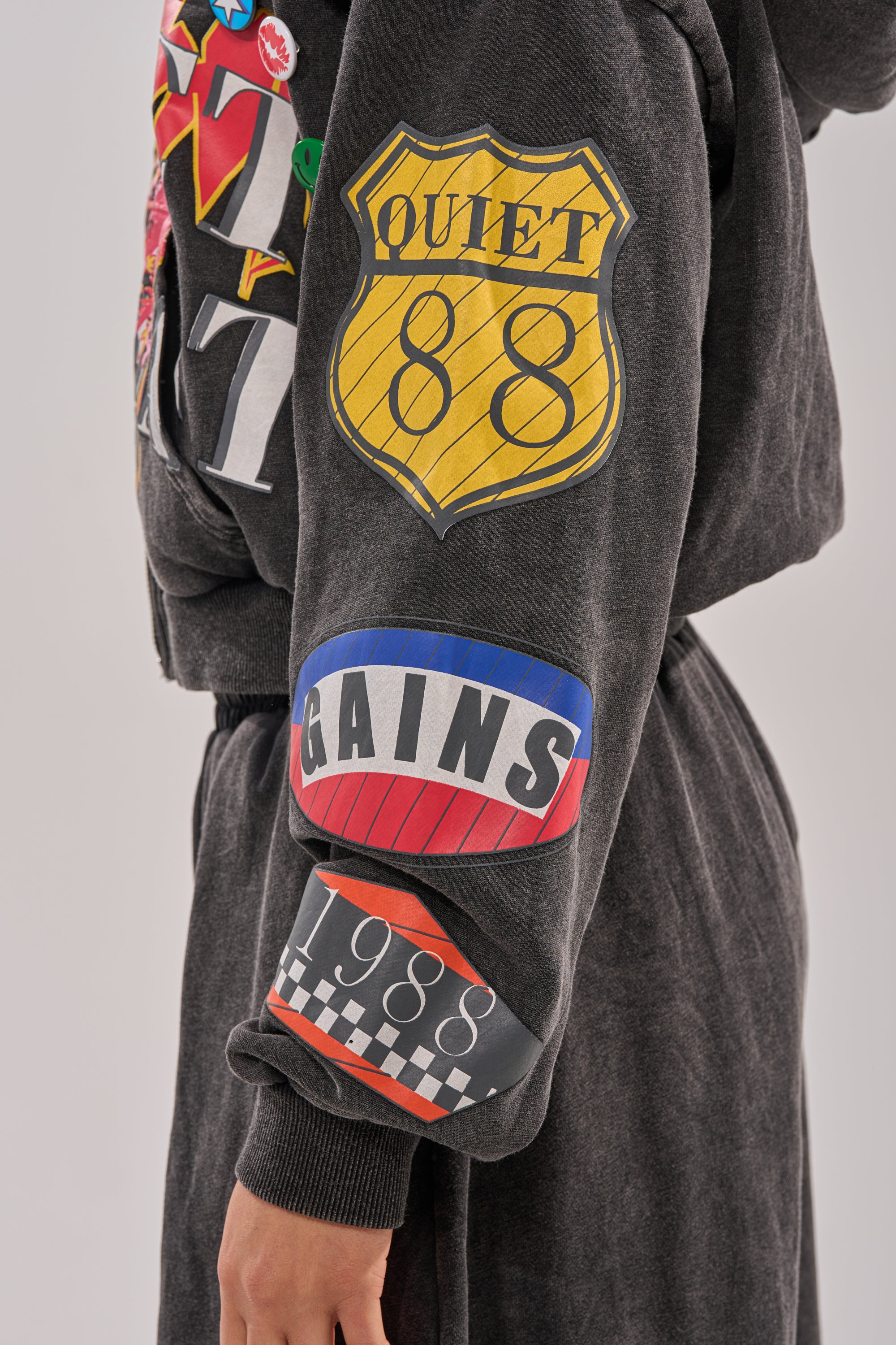 Someone wears the ANOTHER ONE ZIP UP SWEATSHIRT, a dark streetwear hoodie featuring colorful sleeve patches: a yellow "QUIET 88" highway sign, a red-blue "GAINS" patch, and a black-and-white checkered "1988" graphic. 5