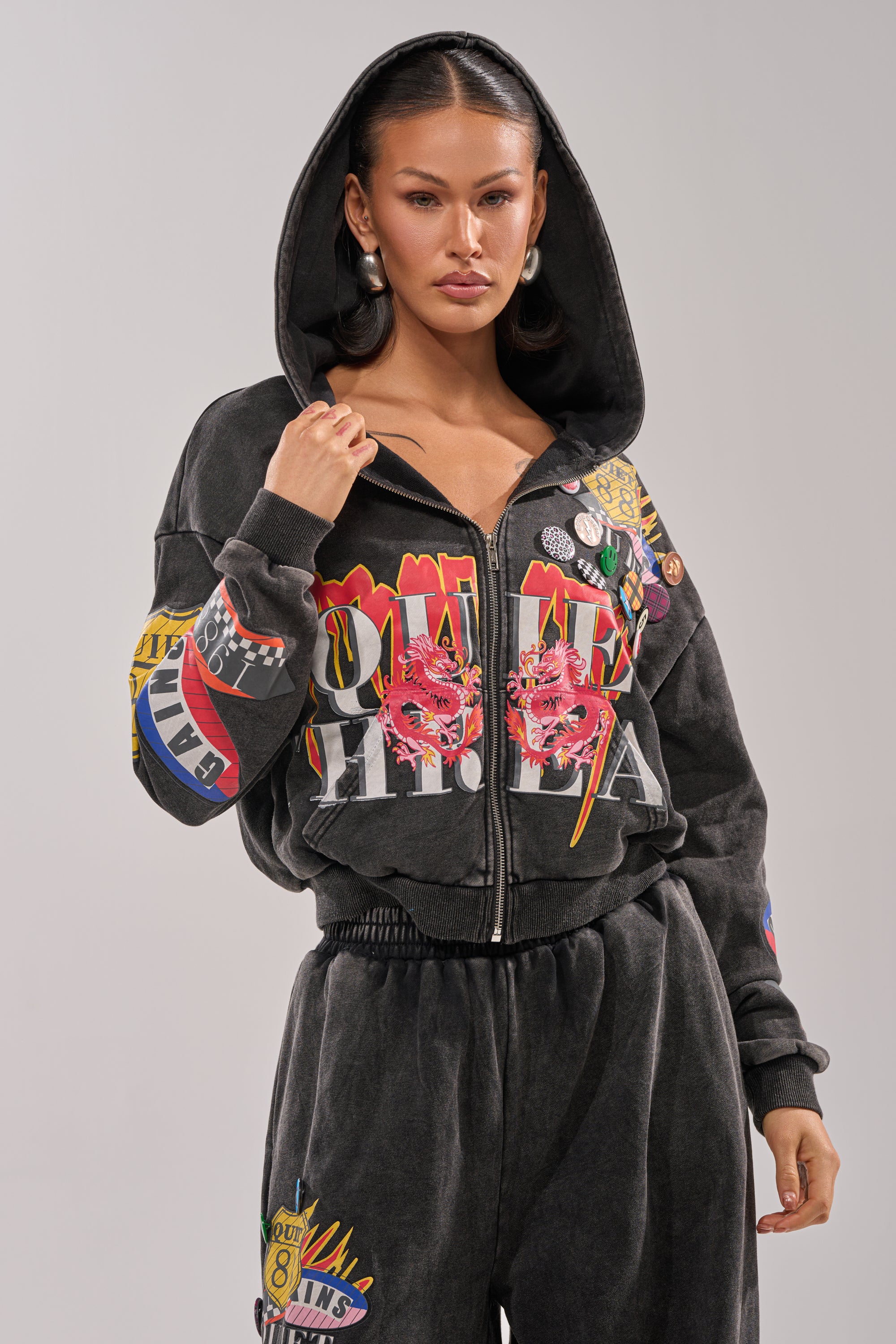A woman exudes streetwear style in the ANOTHER ONE ZIP UP SWEATSHIRT, a black hoodie with vibrant dragon graphics and pins. With her hood up, hands on the zipper, long hair, and hoop earrings, she radiates confidence. 6