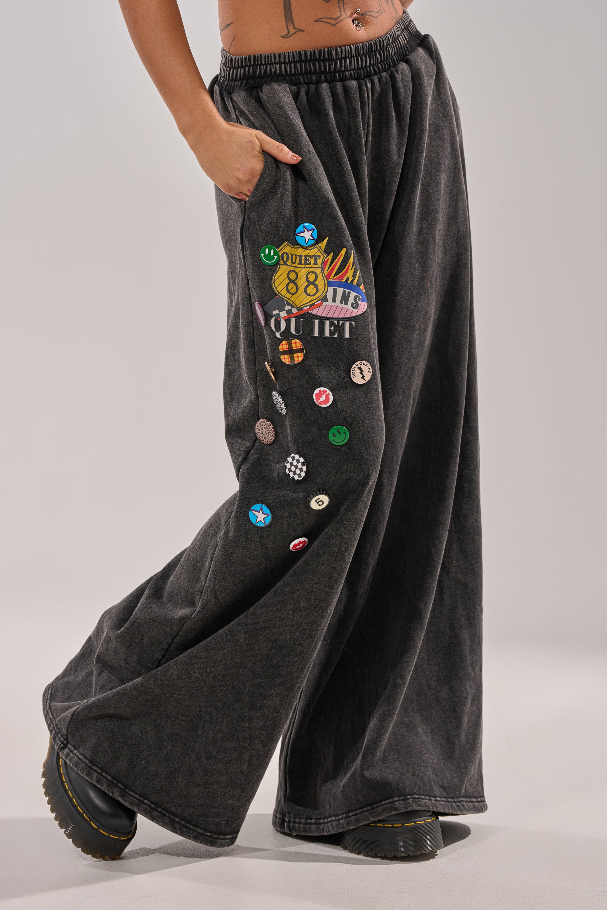 Model wearing the ANOTHER ONE WIDE LEG JOGGER—black streetwear sweatpants with colorful patches and badges on one leg, paired with black platform shoes. One hand in pocket, walking against a plain light gray background. 0