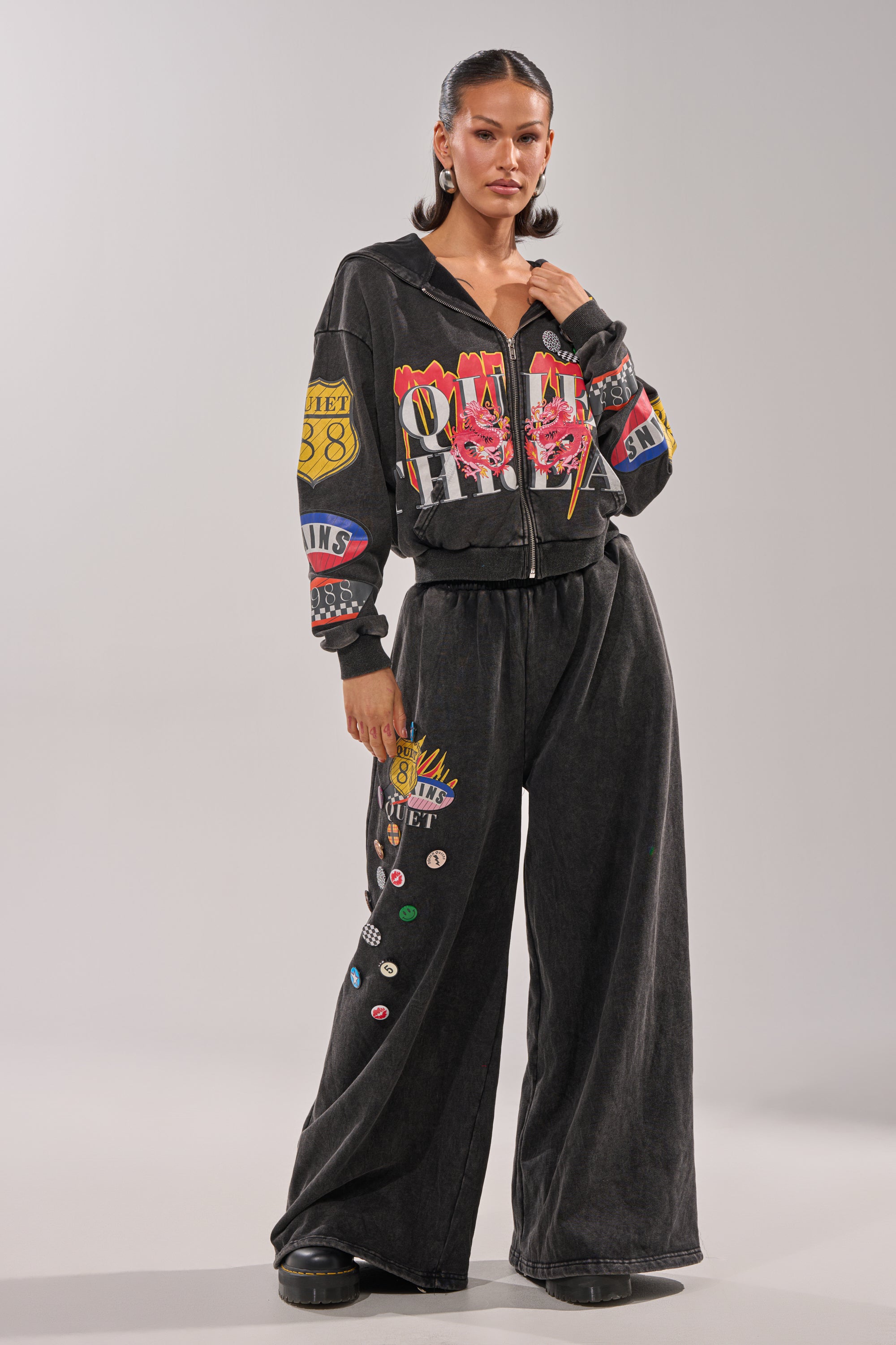 A woman stands confidently in the ANOTHER ONE WIDE LEG JOGGER, featuring colorful graphic patches on black streetwear sweatpants. She has her hair pulled back, wears black platform shoes, and poses against a plain light-colored background. 8