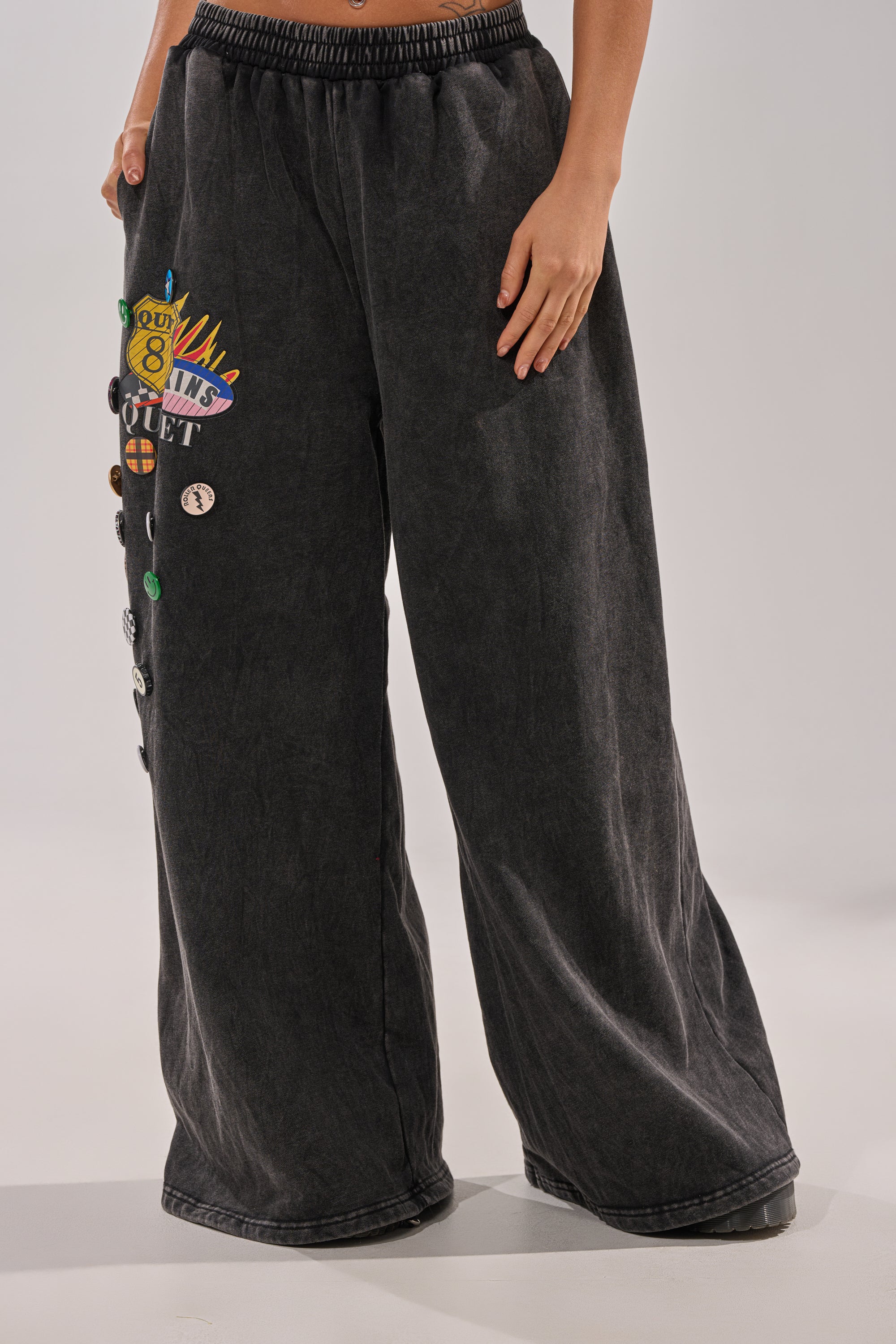 Wearing ANOTHER ONE WIDE LEG JOGGER, loose-fitting black streetwear sweatpants with colorful patches and pins on one side, a person stands against a plain background with their upper body partially out of frame. 1