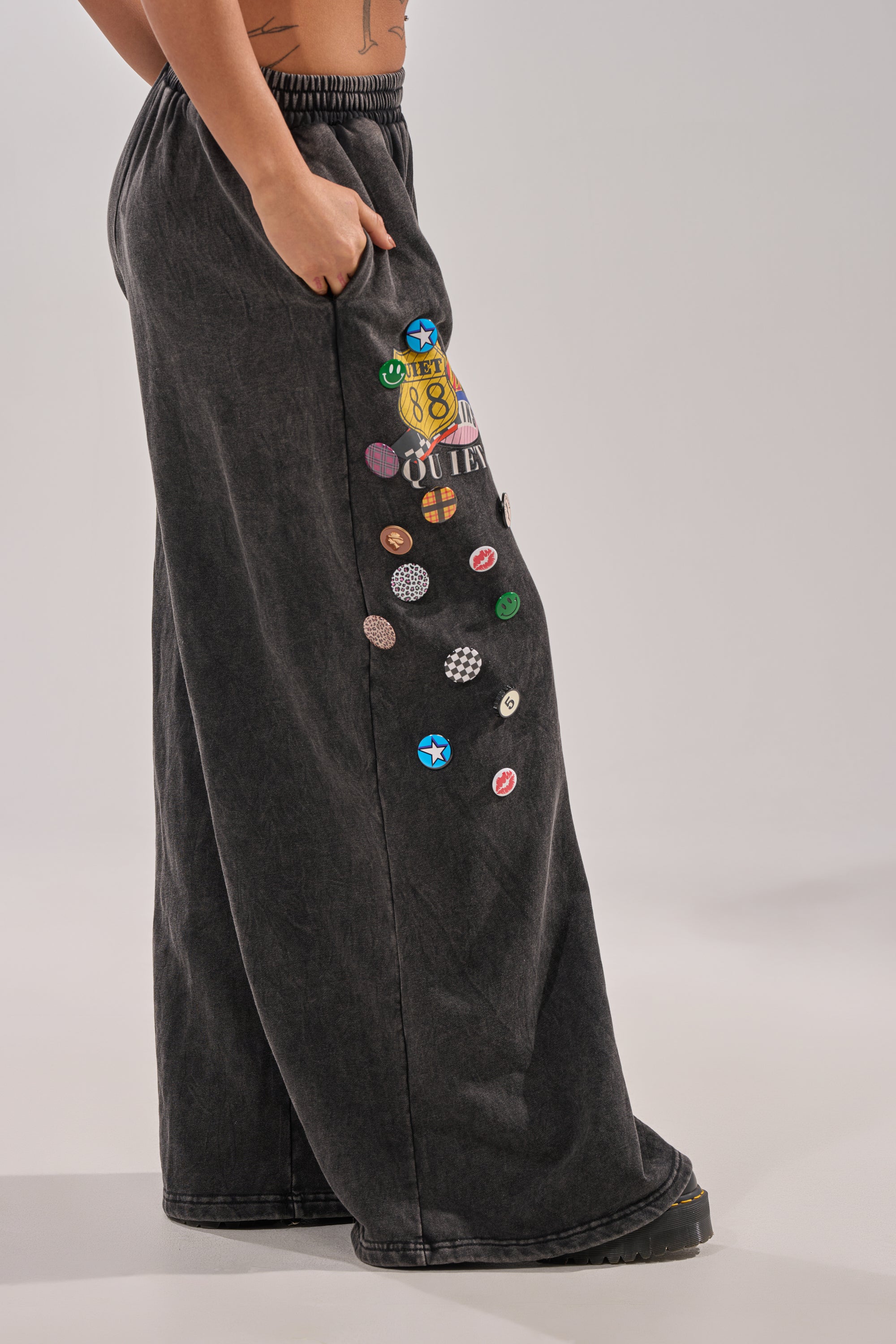 Wearing the ANOTHER ONE WIDE LEG JOGGER, someone stands sideways with one hand in their pocket. The loose-fit dark style with colorful pins and patches gives off a streetwear sweatpants vibe against a plain backdrop. 2