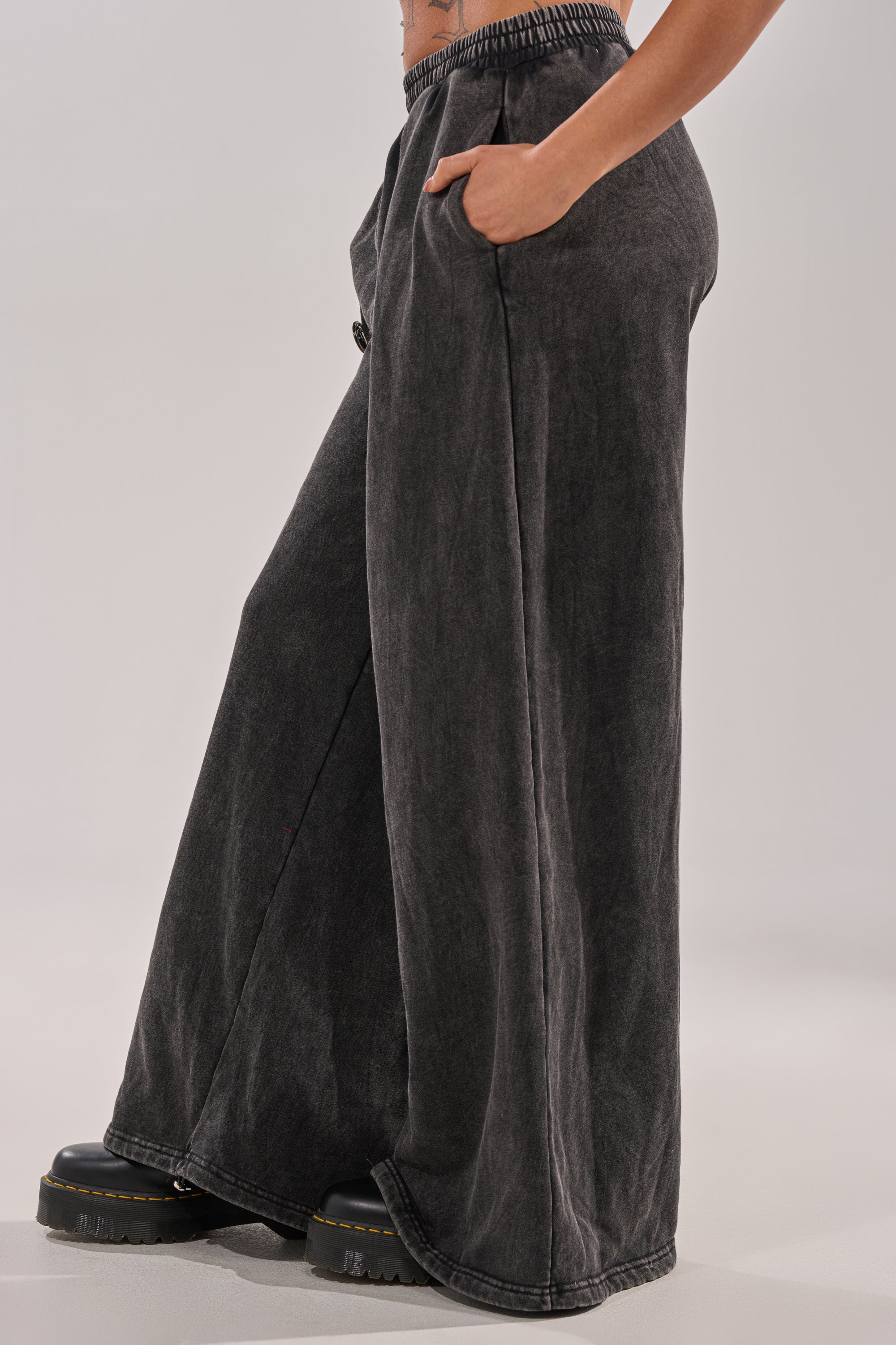 A person stands with one hand in their pocket, wearing the ANOTHER ONE WIDE LEG JOGGER—loose-fit, faded black streetwear pants—and black platform shoes against a plain light background. 3