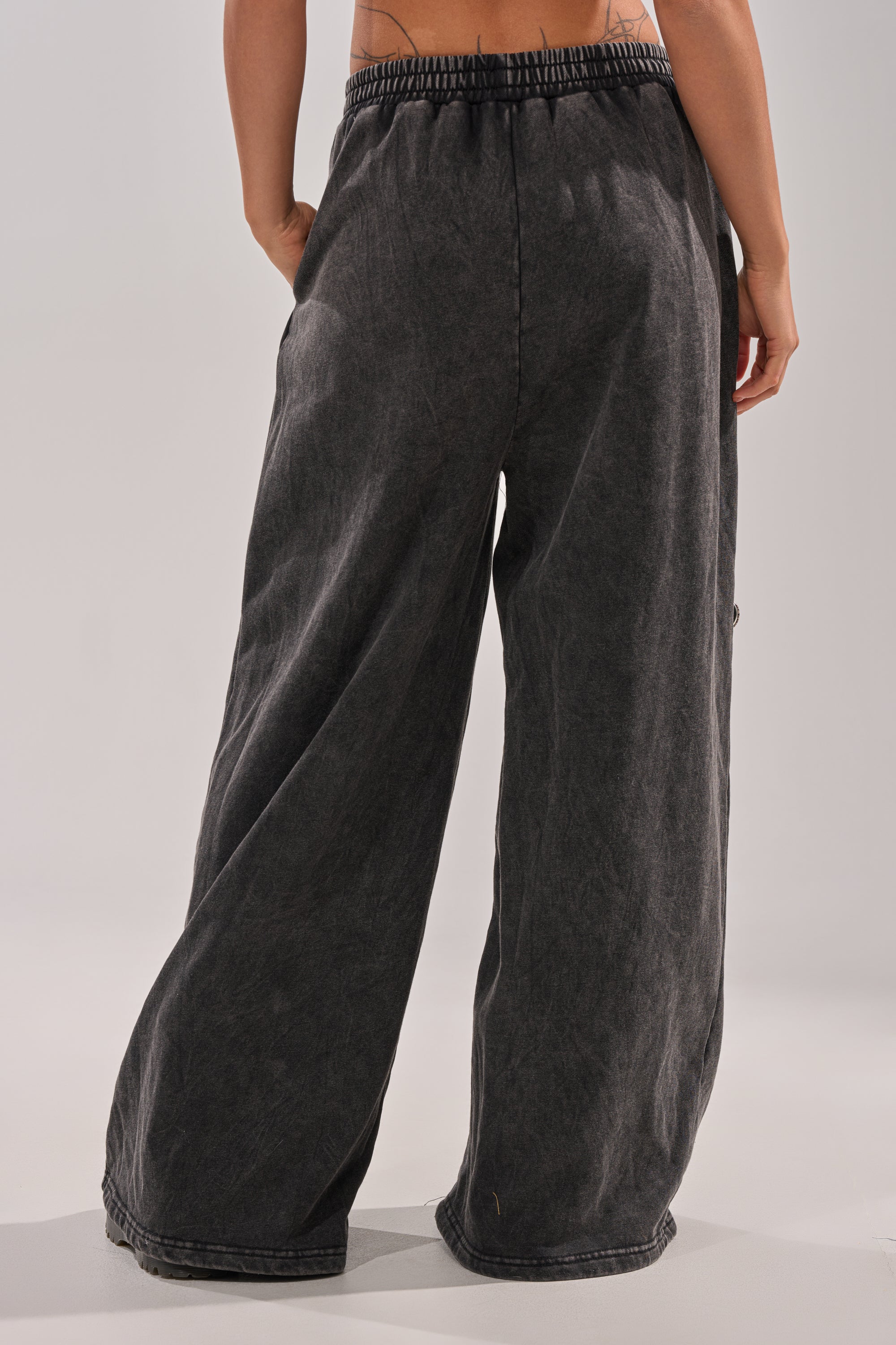 A person stands facing away, showcasing the ANOTHER ONE WIDE LEG JOGGER in dark gray—loose-fitting, high-waisted sweatpants with a wide leg and elastic waistband for a relaxed, casual streetwear look. Only the lower back and legs are visible. 4
