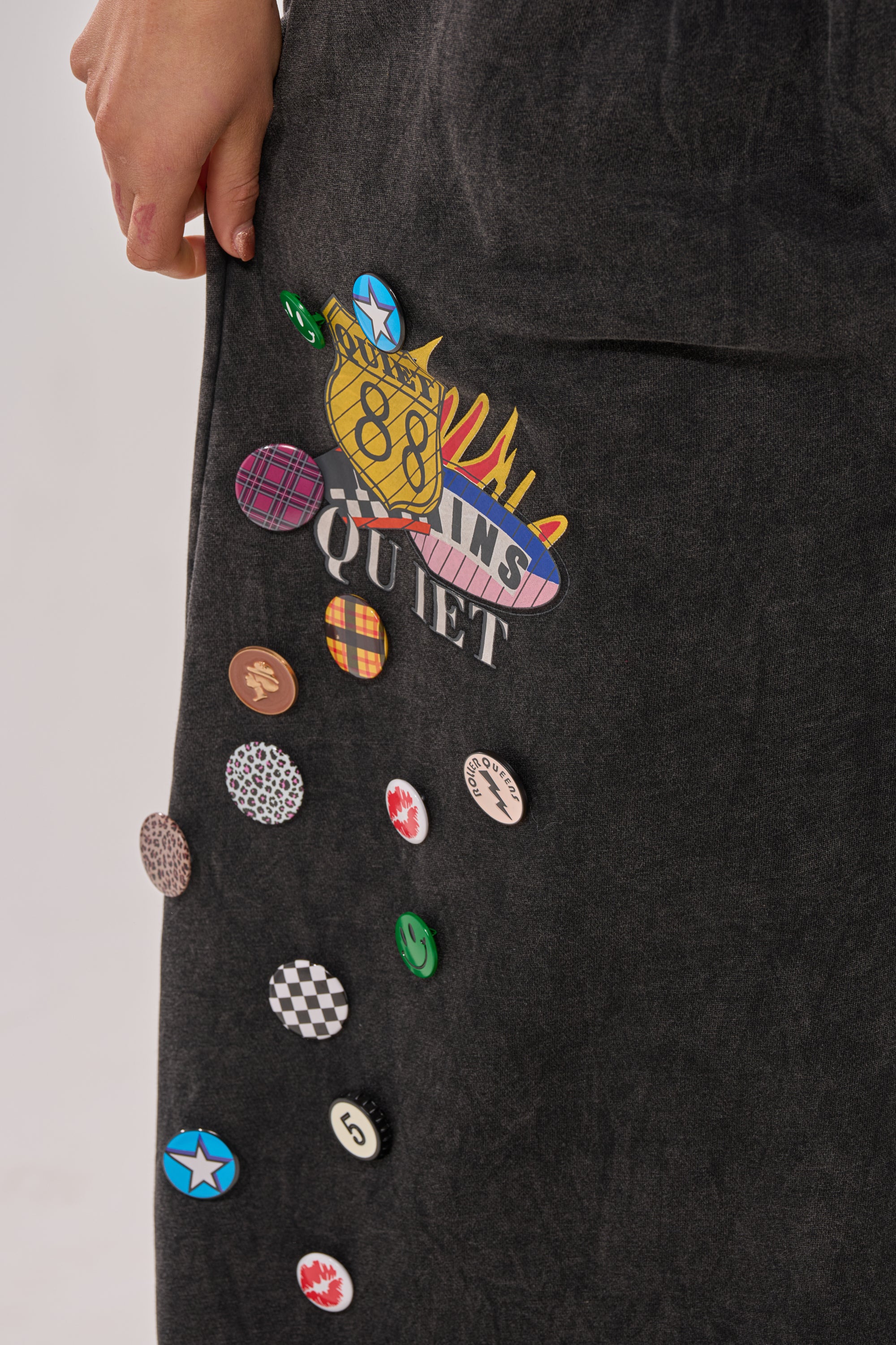A close-up of a hand resting on ANOTHER ONE WIDE LEG JOGGER, adorned with colorful pins and patches like "QUIET," stars, checkerboard, and other vibrant designs. 6