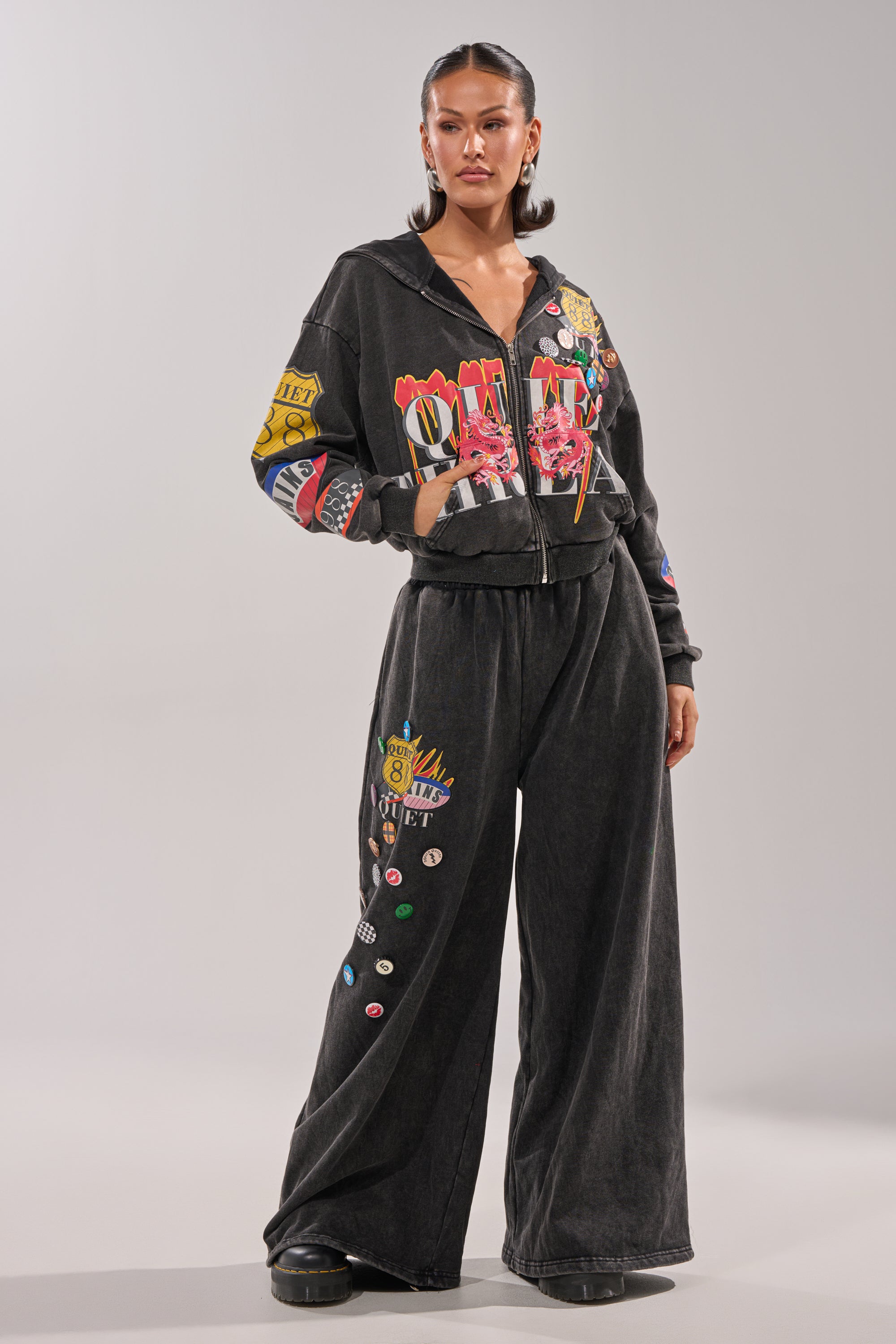 A woman stands confidently in the ANOTHER ONE WIDE LEG JOGGER and matching zip-up hoodie, both featuring colorful graphic patches. With slicked-back hair and black platform shoes, she completes her bold streetwear look. 7