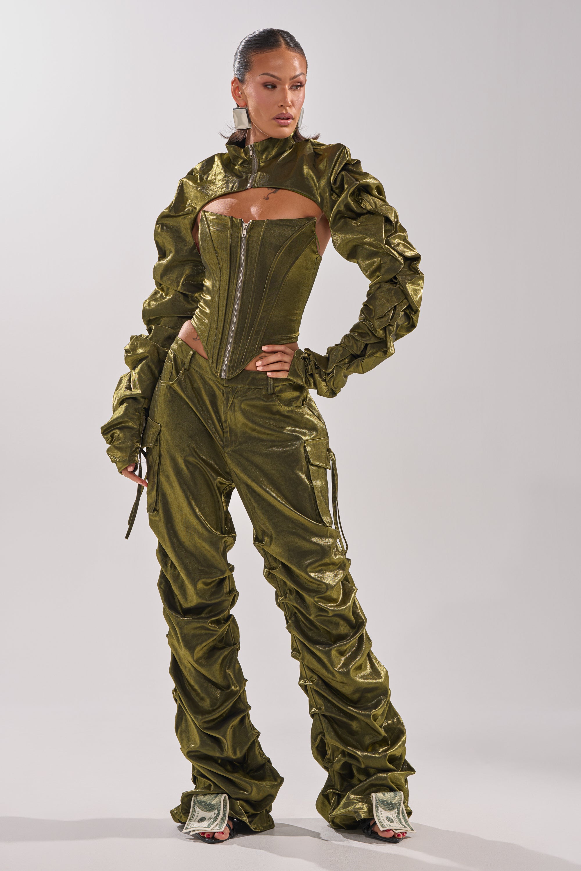 A woman models the LUCKY ONE STACKED TROUSER: olive green, shiny, ruched pants with a stacked silhouette. She pairs them with a matching corset-style zip-up top and heels, accessorized with hoop earrings. Her hair is slicked back; background is light gray.