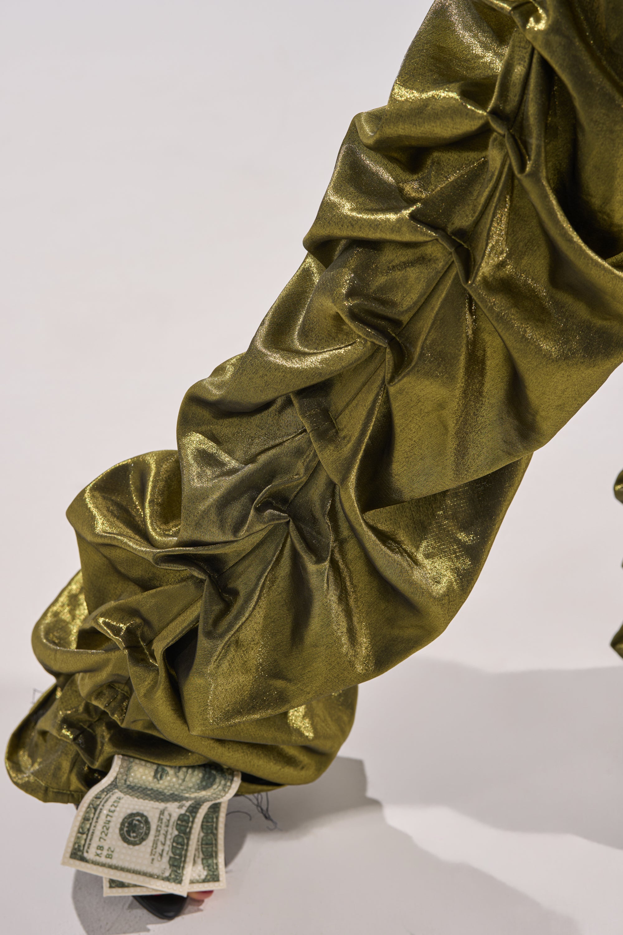 The LUCKY ONE STACKED TROUSER in shiny, ruched metallic olive-green fabric drapes over a white surface, partially revealing a few U.S. dollar bills tucked underneath.
