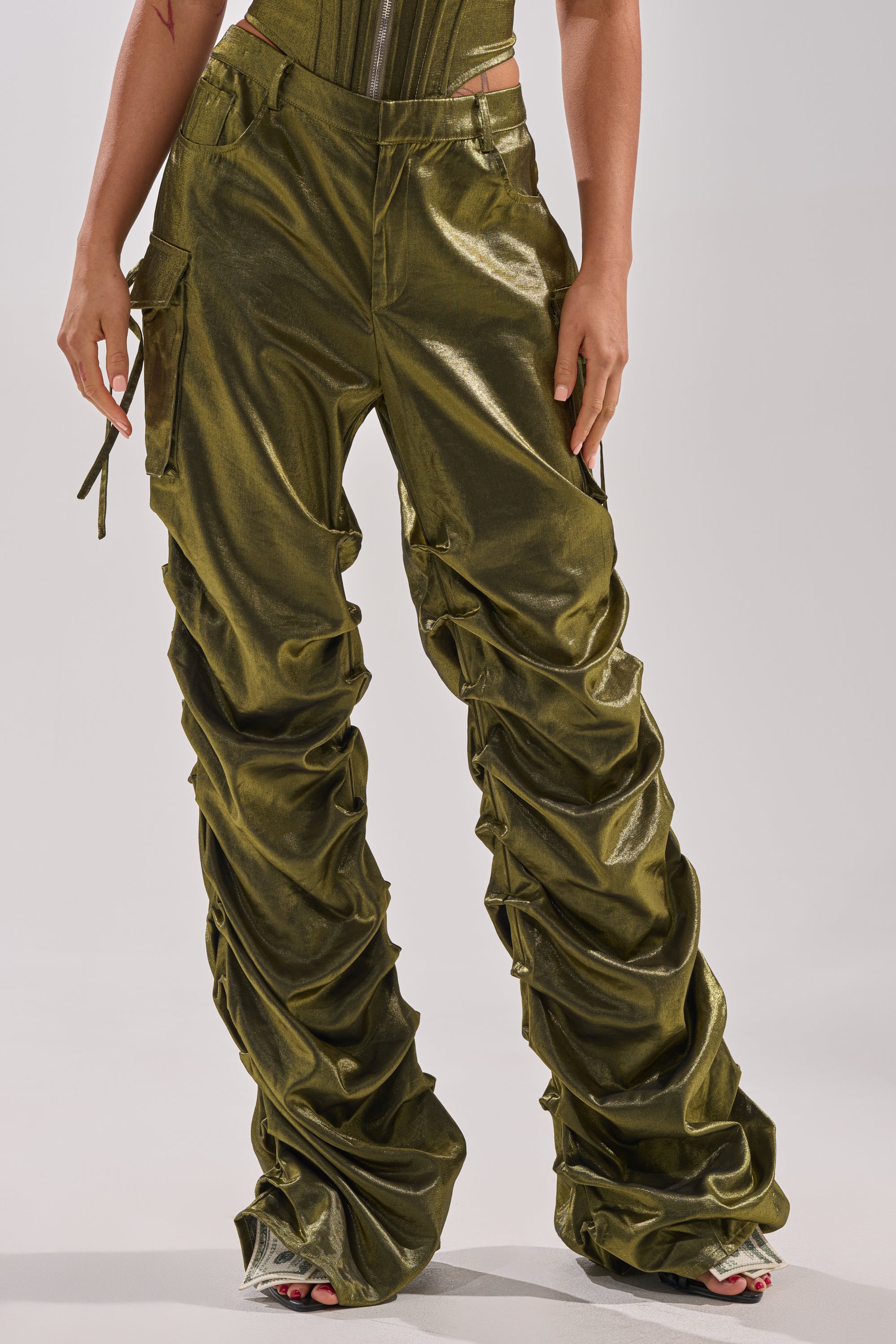 Someone stands with hands at their sides, wearing shiny olive green LUCKY ONE STACKED TROUSER by AKIRA Label—these straight-leg pants feature cargo-style pockets, gathered ruched details, and wide legs that cover most of the feet.
