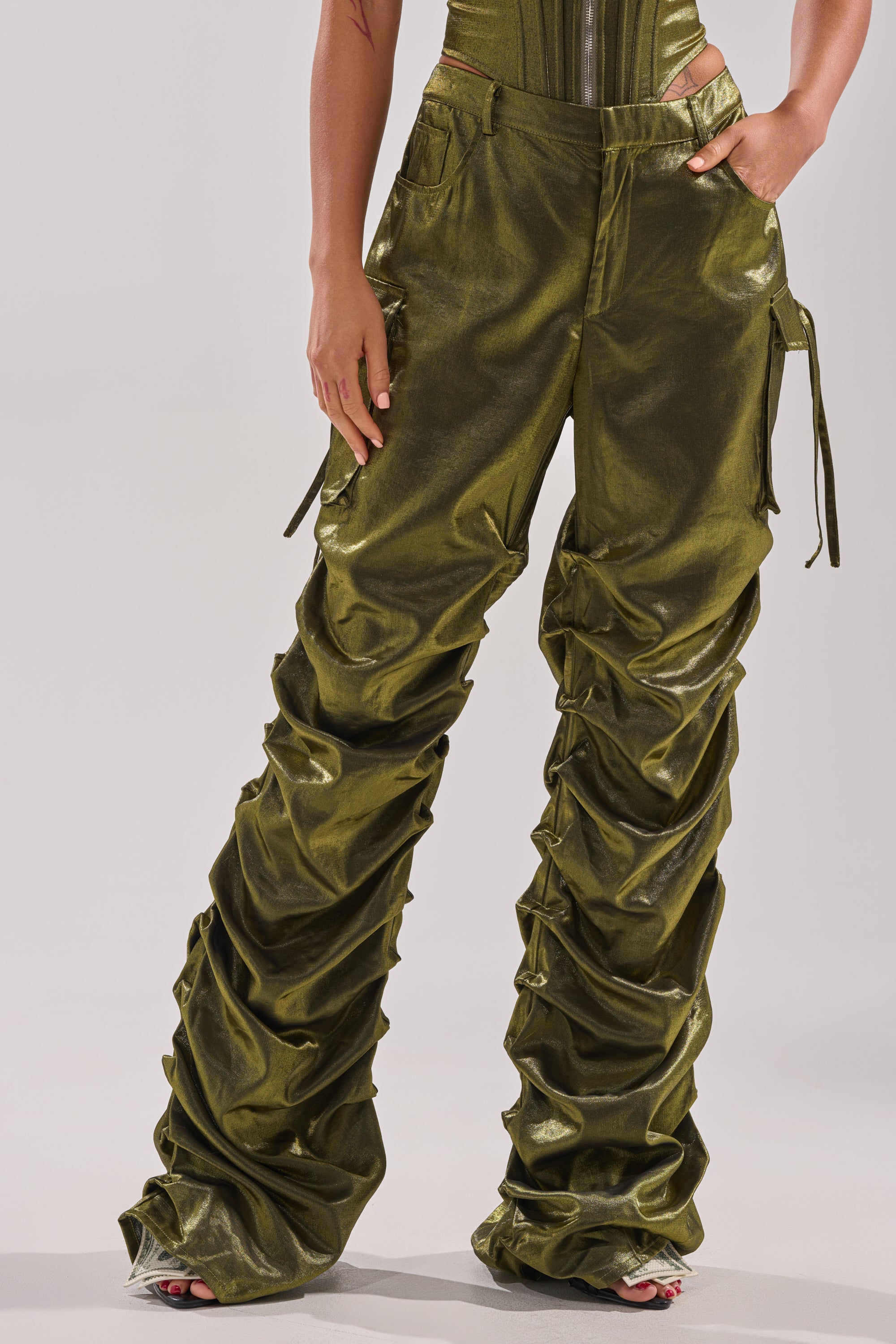 A person wears shiny, olive green LUCKY ONE STACKED TROUSER featuring ruched straight-leg pants, cargo-style pockets, gathered detailing, tie waist accents, and a matching fitted top.