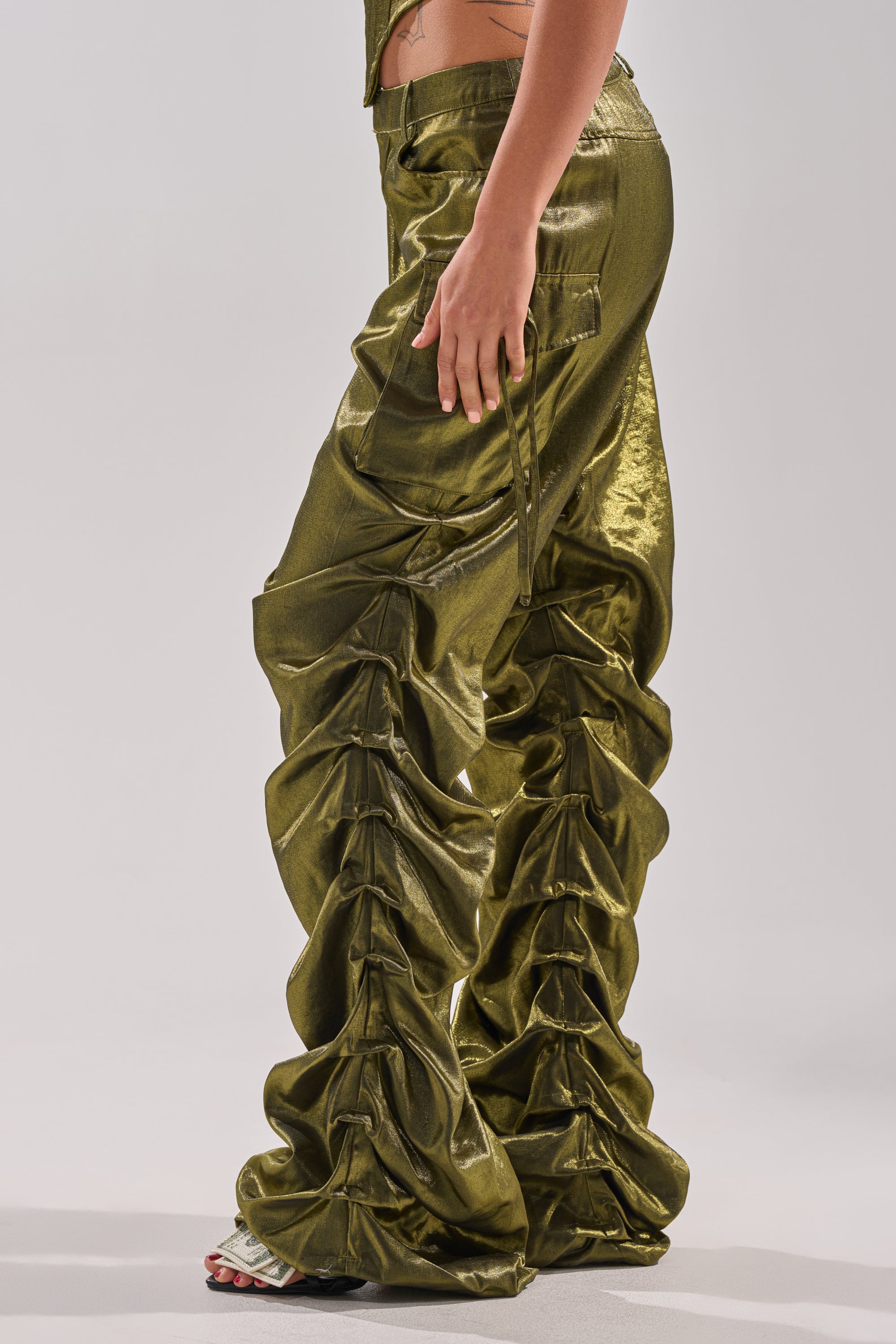 A person stands sideways on a light background, showcasing only their lower torso and legs in the shiny, metallic olive-green LUCKY ONE STACKED TROUSER with ruched, gathered detailing along the legs.