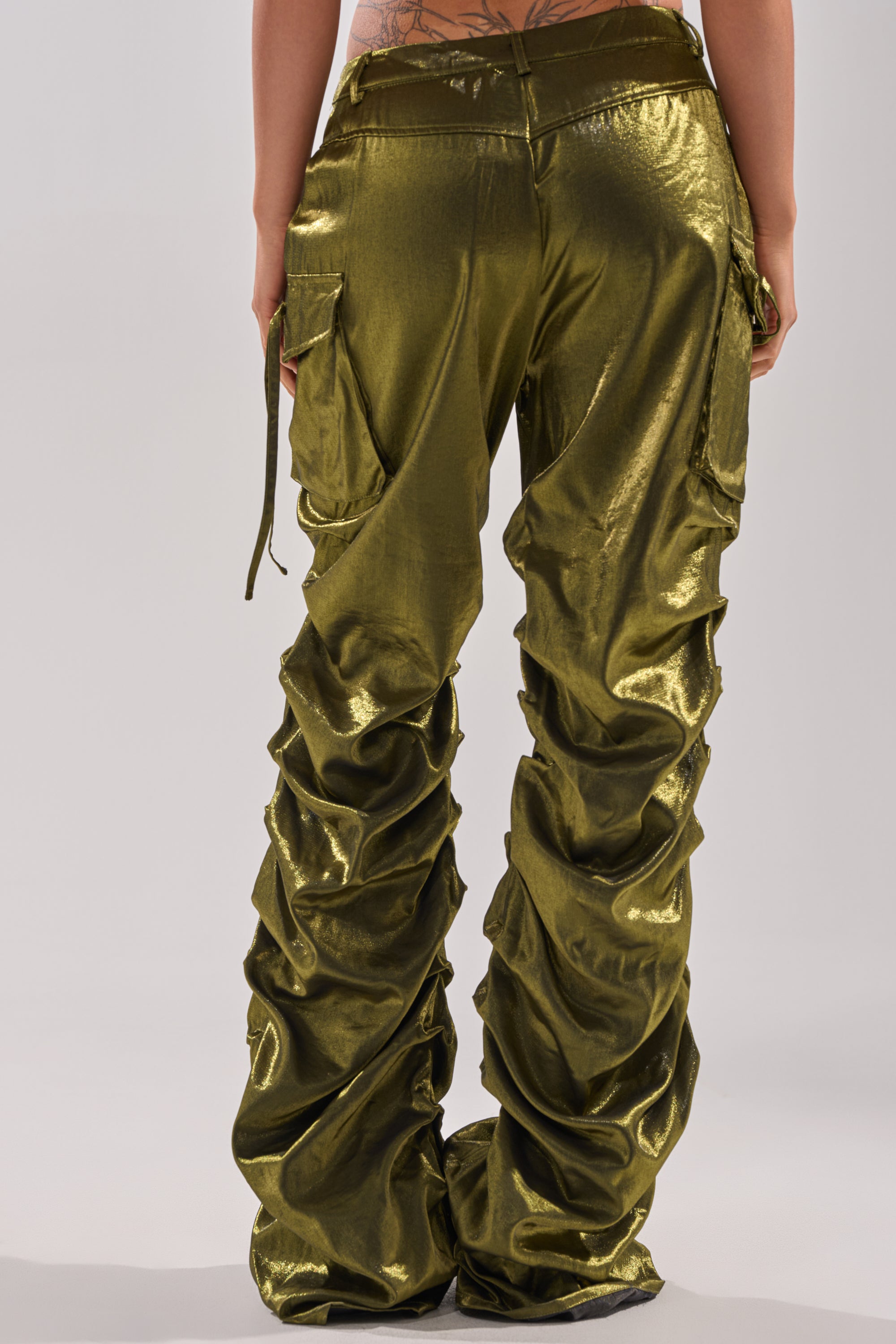 The LUCKY ONE STACKED TROUSER features shiny olive green fabric, ruched detailing along the legs, cargo pockets, and a modern stacked silhouette—seen from the back for a textured, statement look.