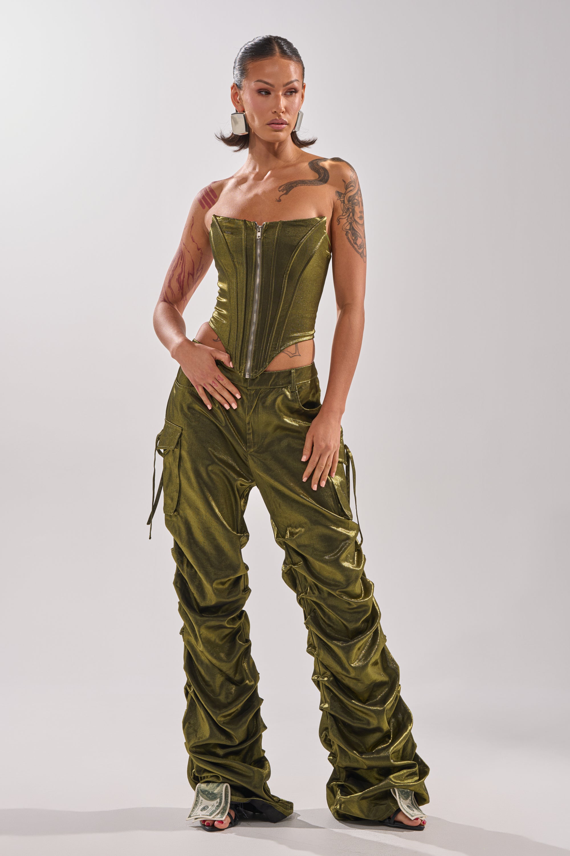 A woman with tattoos and styled dark hair stands confidently against a plain background, wearing a strapless olive green corset top and LUCKY ONE STACKED TROUSER paired with matching ruched pants.