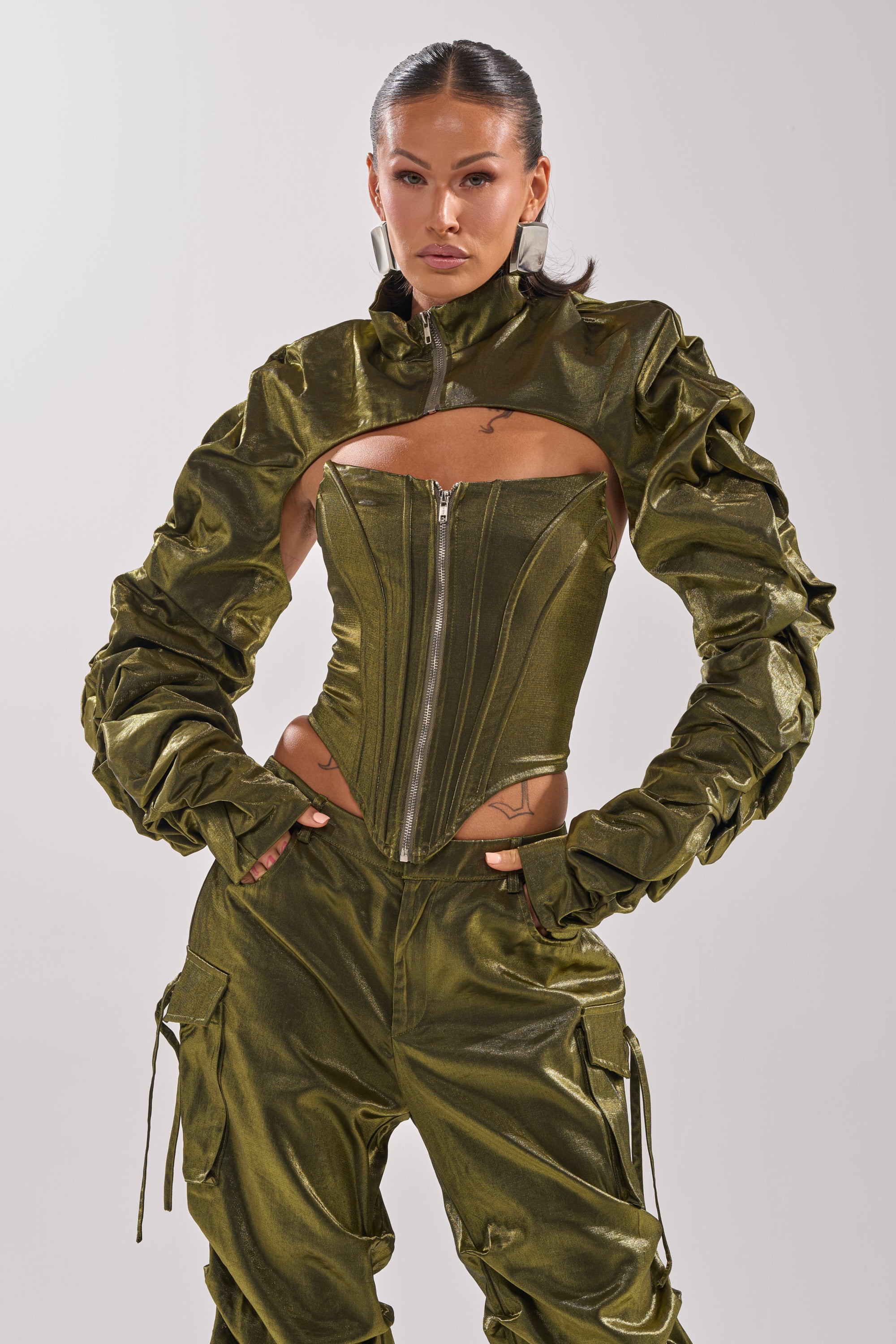 A woman stands confidently in the LUCKY ONE BOLERO, a shiny olive green piece featuring ruched sleeves and a zip-up corset top. She accessorizes with hoop earrings, slicked-back hair, and poses with hands on hips against a plain background. 1