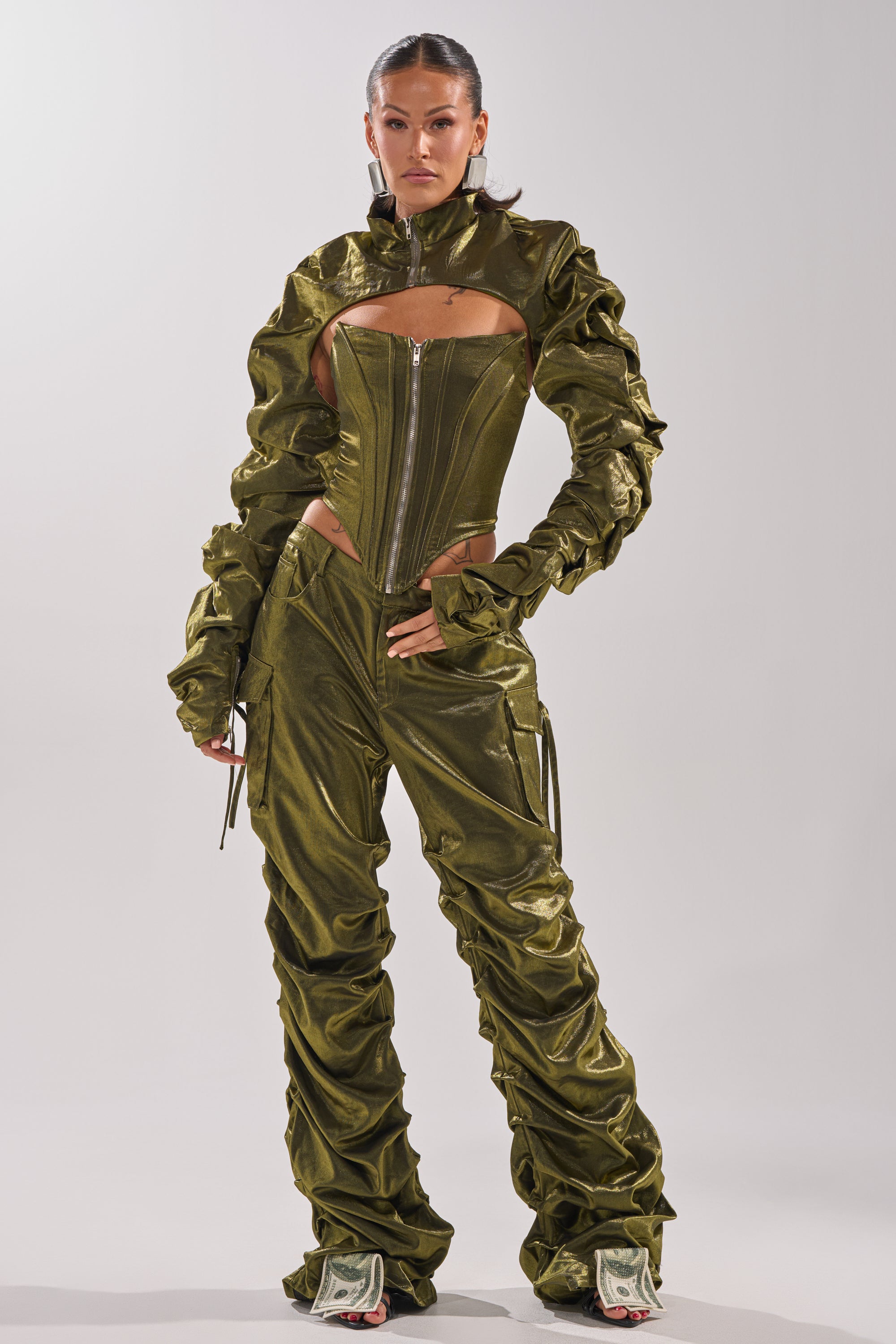 A woman exudes confidence in an olive green, textured pantsuit with ruched details and a zip-up corset top, styled with the LUCKY ONE BOLERO and standout dollar bill print shoes. Her hair is slicked back in a high ponytail. 5