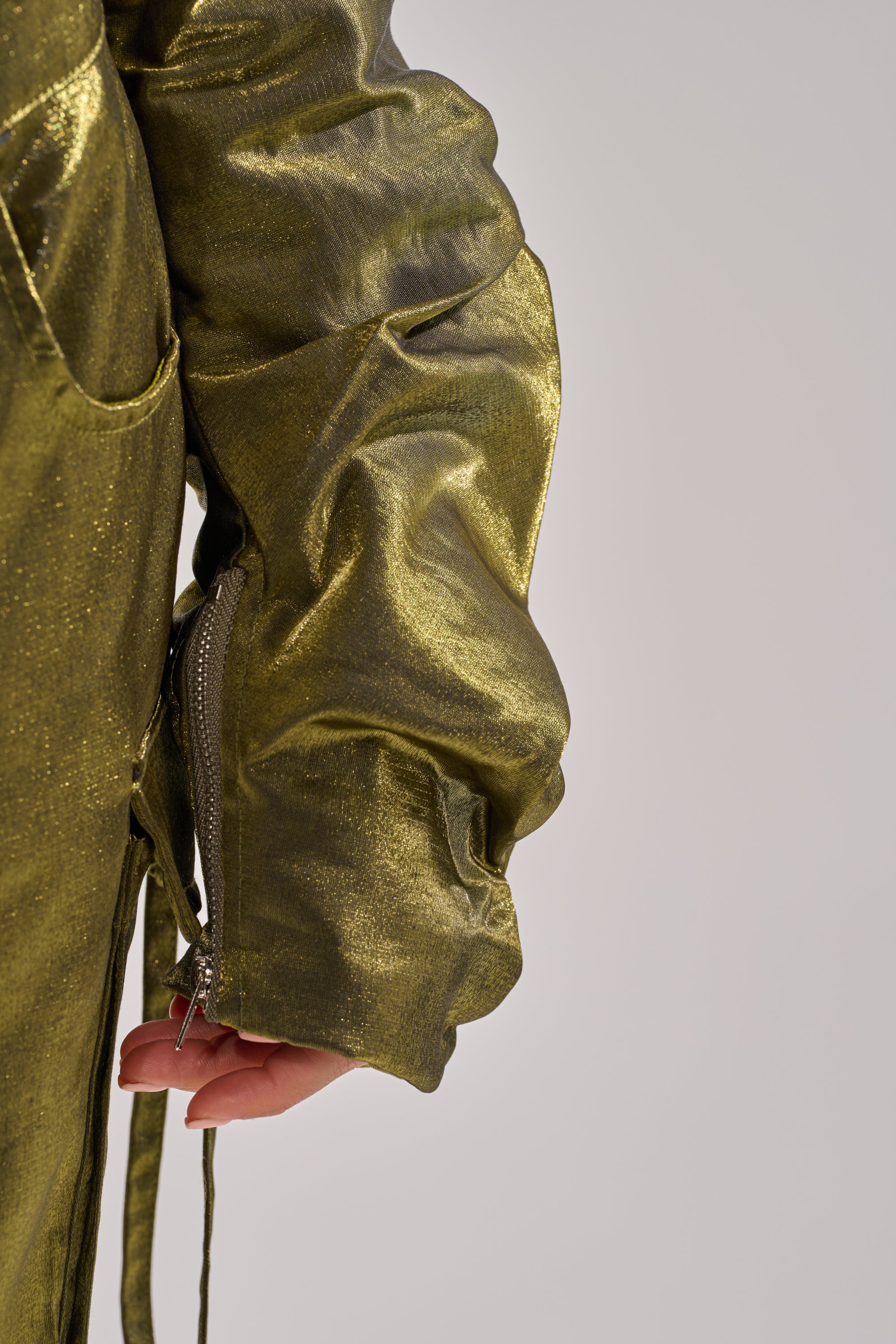 A close-up of a hand partially tucked into the pocket of the LUCKY ONE BOLERO, a shiny metallic gold ruched cropped jacket with a visible sleeve zipper, set against a plain light background. 8