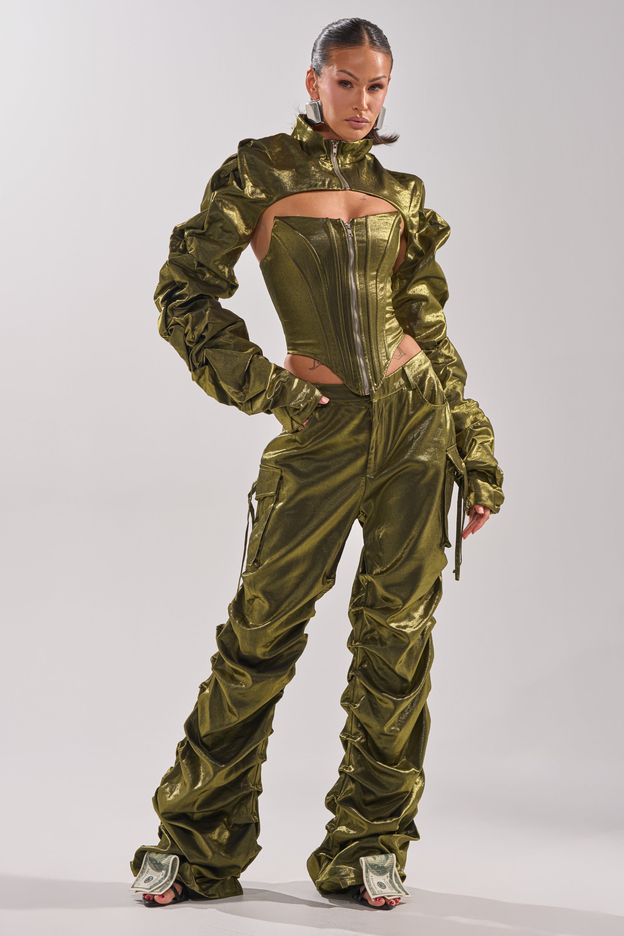 A woman poses confidently in the AKIRA Label LUCKY ONE CORSET, a strapless olive green top styled with ruched sleeves, matching pants, and heels adorned with dollar bills. Her hair is slicked back.