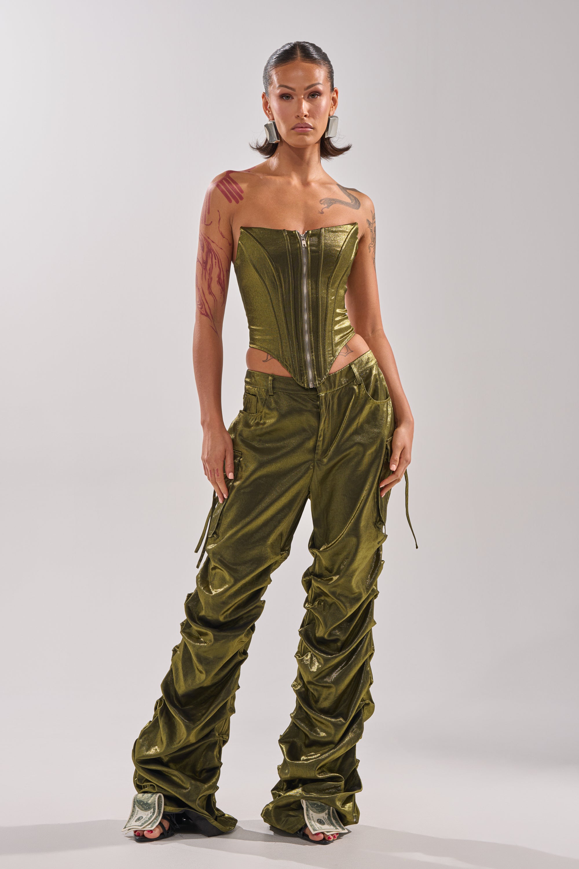 A woman with slicked-back hair and arm tattoos stands confidently against a light background wearing the LUCKY ONE CORSET, an olive green strapless top paired with matching wide-leg ruched pants.