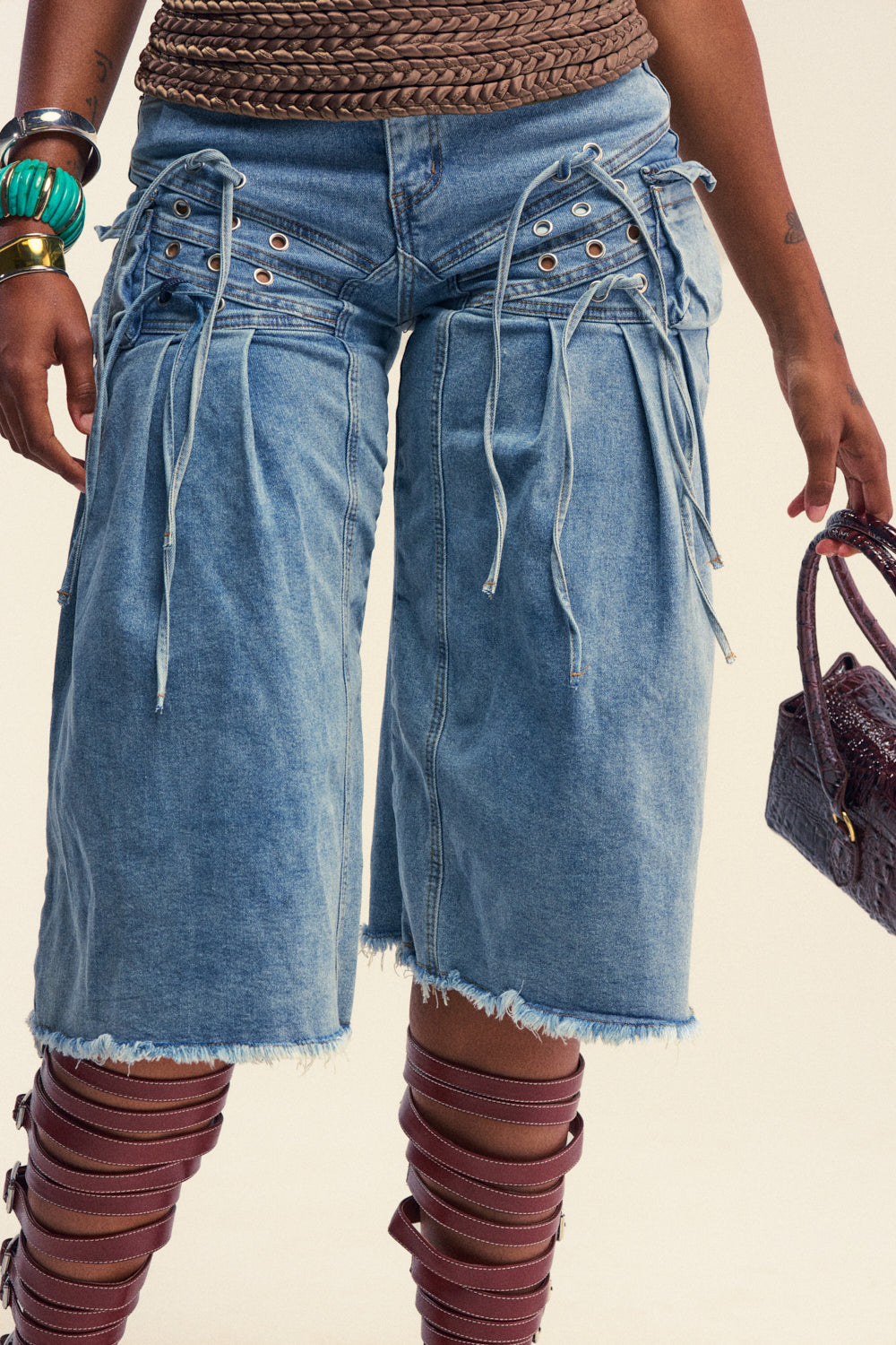 Wearing the BABY GIRL DENIM GAUCHO SHORT in knee-length light blue denim, featuring lace-up accents and frayed hems, styled with bangles, a woven belt, a textured handbag, and maroon strappy leg wraps.