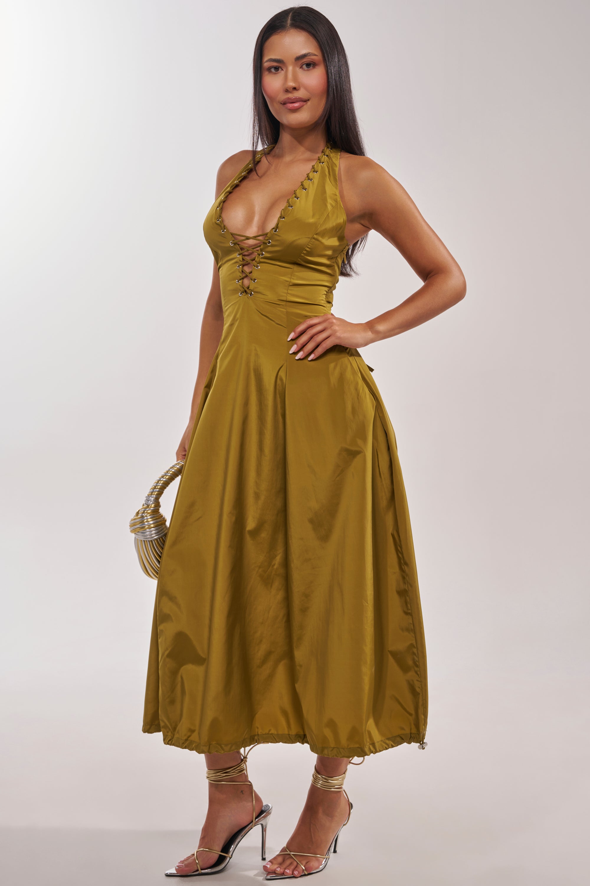 A woman stands confidently in the SOMETHING LIGHT NYLON MAXI DRESS—a sleeveless halter-neck maxi with a gold lace-up detail and mid-length skirt. She completes her look with a small handbag and strappy high-heeled sandals. 3