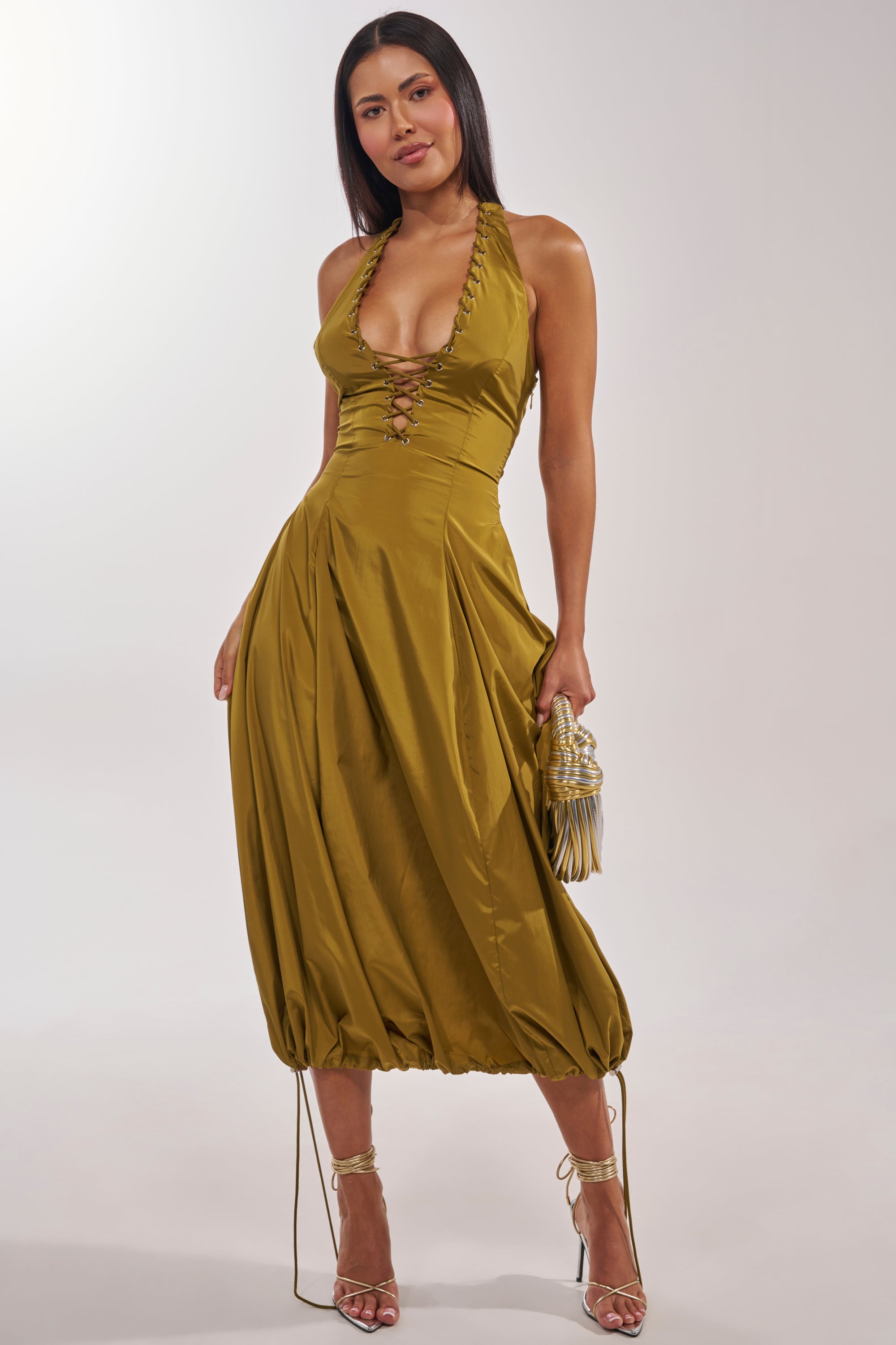 A woman models the SOMETHING LIGHT NYLON MAXI DRESS in shiny olive with a deep neckline and lace-up front, holding a small ruched handbag. She pairs it with strappy high-heeled sandals and wears her straight, dark hair down. 4