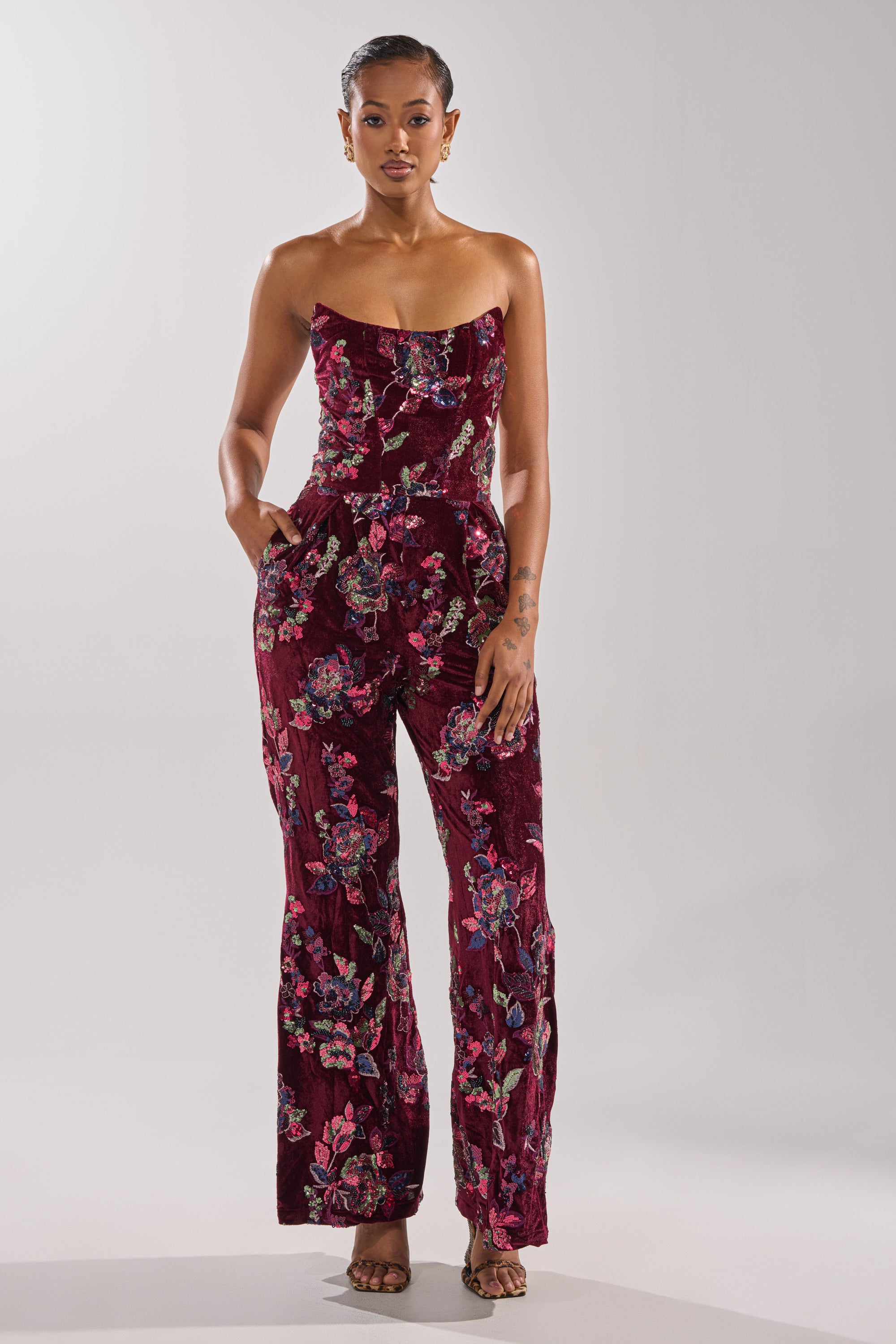 A woman with short hair stands against a light gray background wearing the IN YOUR DREAMS VELVET JUMPSUIT—a strapless burgundy velvet jumpsuit with a colorful floral pattern, hands in her pockets and paired with open-toed heels.