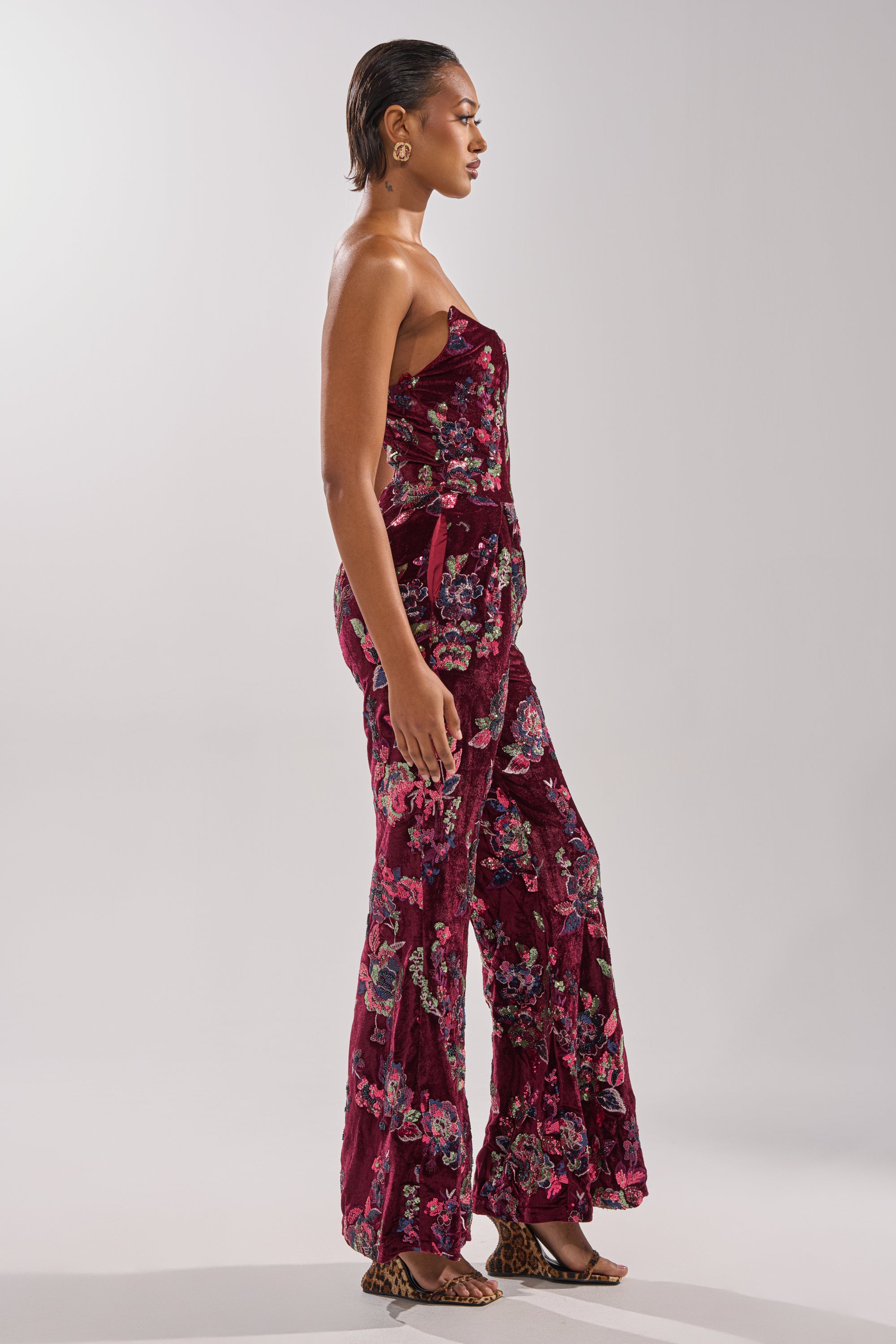 A woman stands in profile wearing the IN YOUR DREAMS VELVET JUMPSUIT, which is strapless with sequin floral embroidery and flared legs. She has short, slicked-back hair, hoop earrings, open-toe sandals, and poses against a light plain background.