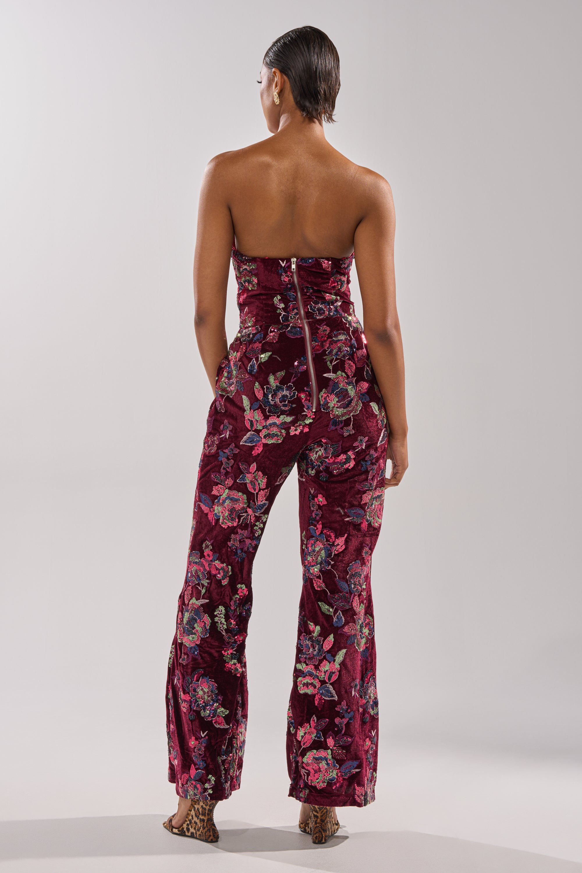 Someone stands with their back to the camera in the IN YOUR DREAMS VELVET JUMPSUIT, featuring strapless burgundy velvet and bold sequin floral embroidery. Their hair is slicked back, paired with open-toed heels, against a plain light gray background.