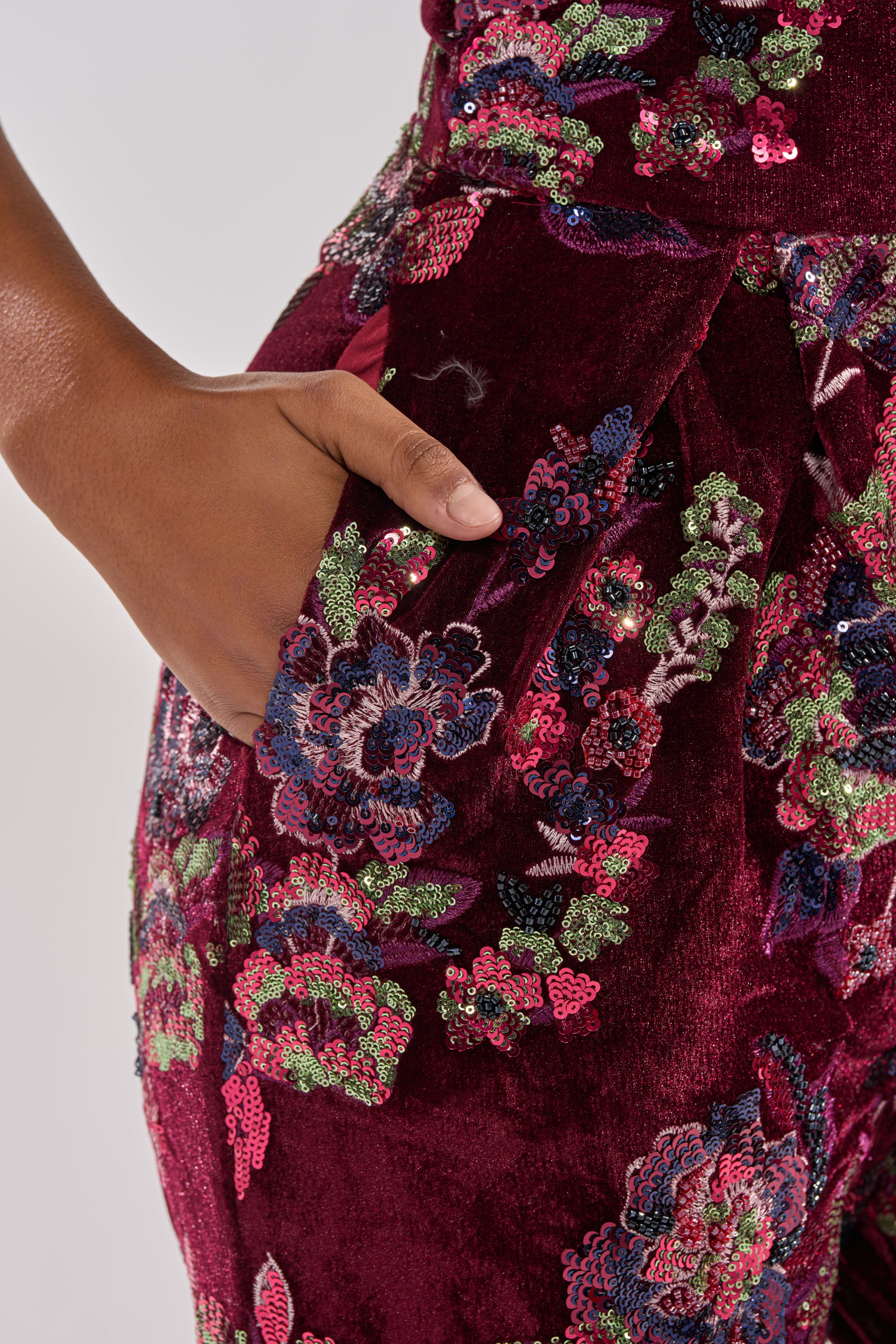A close-up of a hand in the pocket of the IN YOUR DREAMS VELVET JUMPSUIT, featuring a burgundy velvet fabric with vibrant sequin floral embroidery. The background is plain and light-colored.