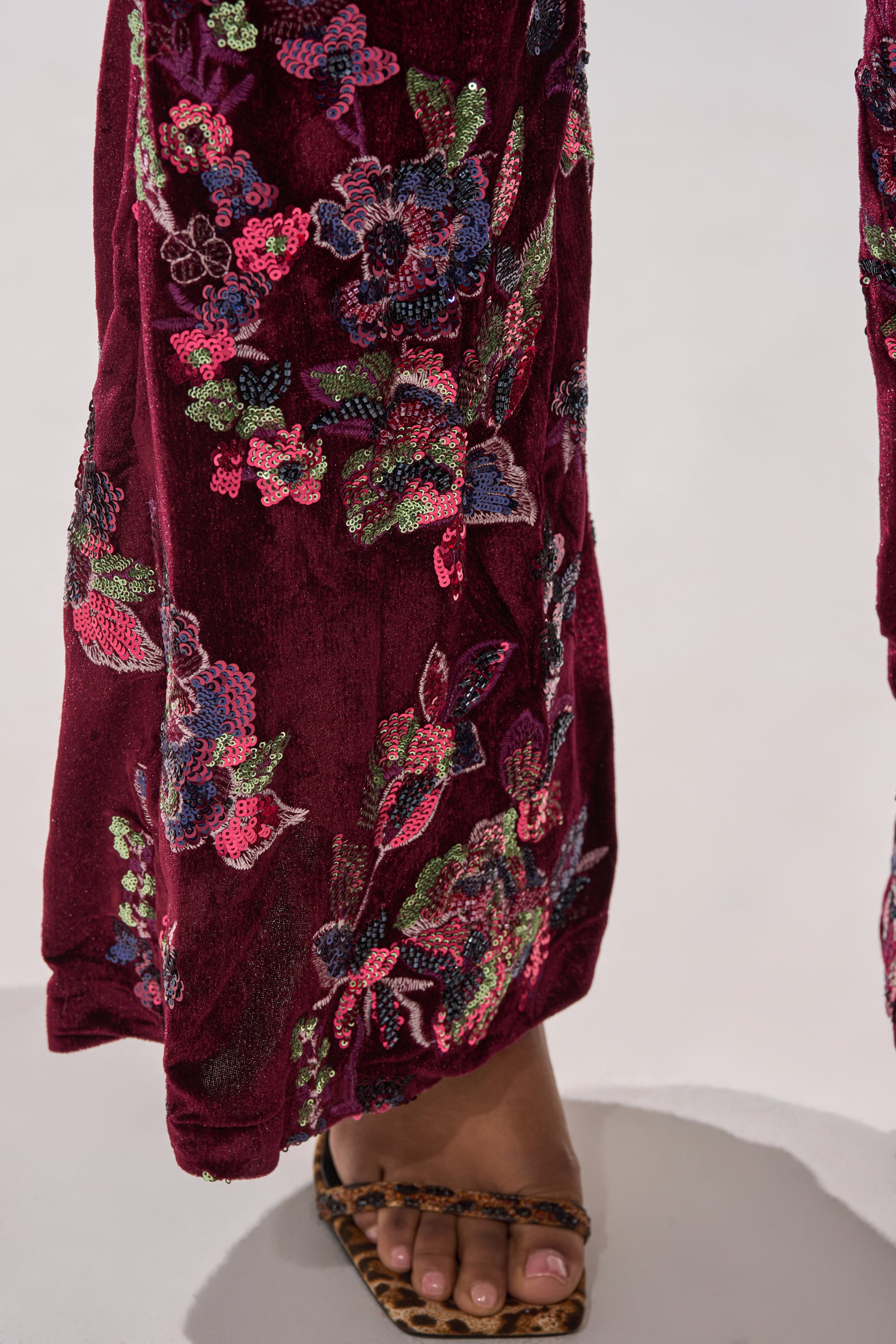 A close-up of a lower leg in the IN YOUR DREAMS VELVET JUMPSUIT shows burgundy velvet pants with colorful sequin floral embroidery, paired with an open-toe sandal against a light background.