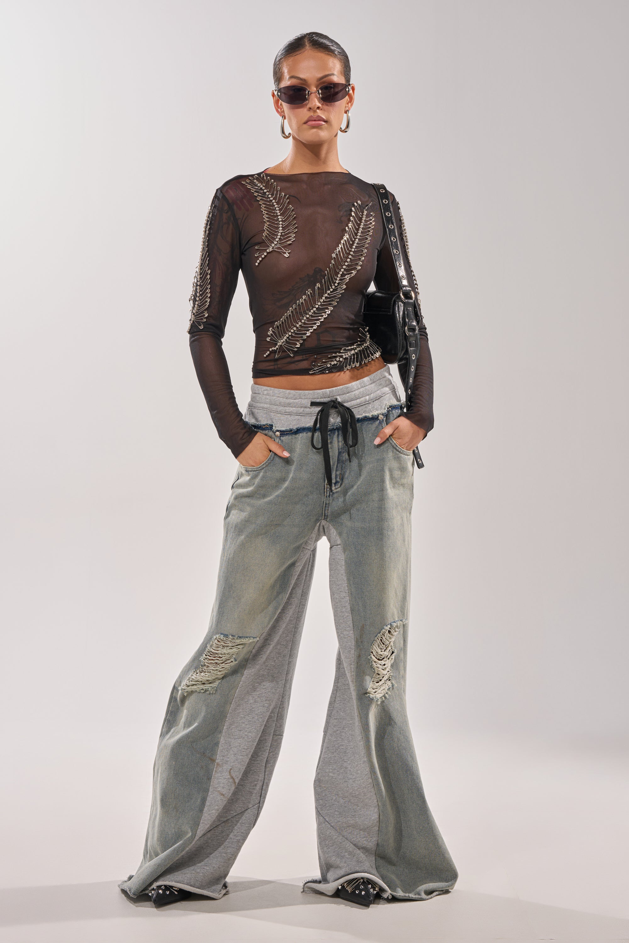 A person stands against a plain background, hands in pockets, wearing sunglasses, a sheer black top with metallic feather designs, pointed shoes, and the COMFY WIDE LEG DENIM JOGGER featuring flared legs and gray panels. 0