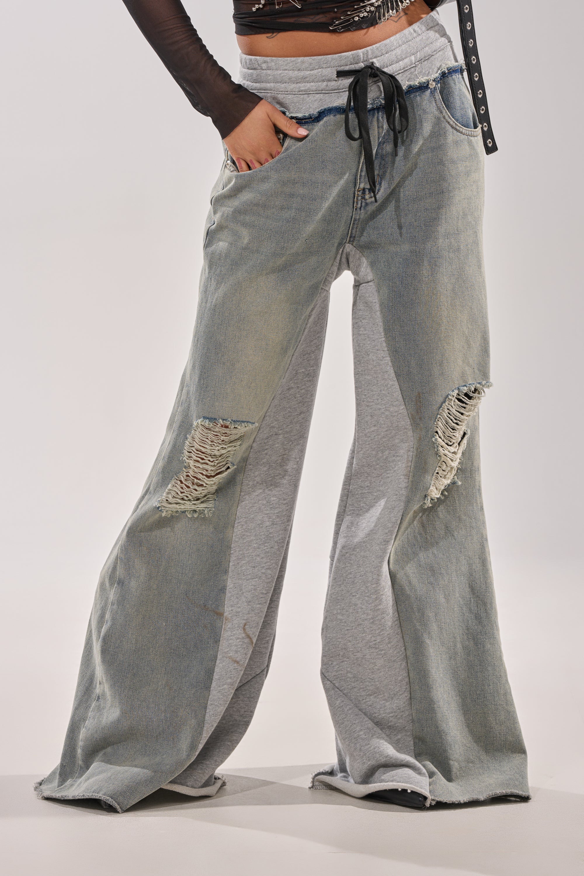 Model wears the COMFY WIDE LEG DENIM JOGGER featuring flared legs, gray sweatpant inserts, distressed thigh patches, black belt, and a cropped top—perfect for standout street-style against a plain background. 1