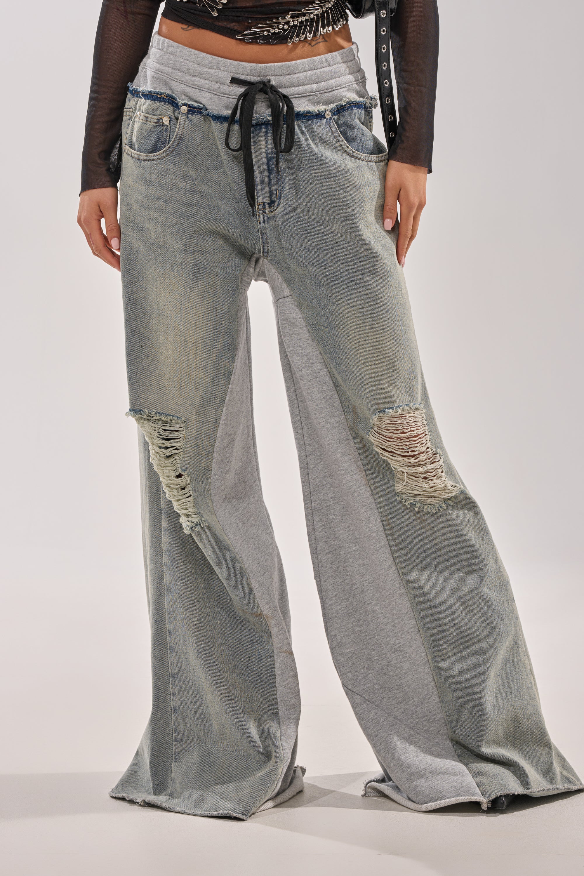 Wearing the COMFY WIDE LEG DENIM JOGGER, featuring wide legs, light gray inserts, ripped knees, a black drawstring waist, and a bold two-tone street-style design on a neutral base. 2