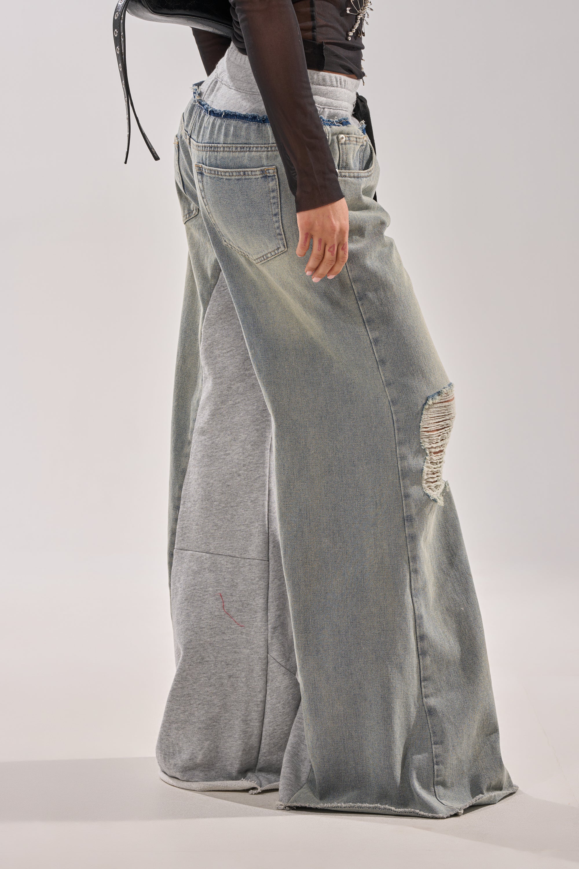 Someone wears the COMFY WIDE LEG DENIM JOGGER layered over gray sweatpants with a ripped right knee, standing against a plain light background. 3