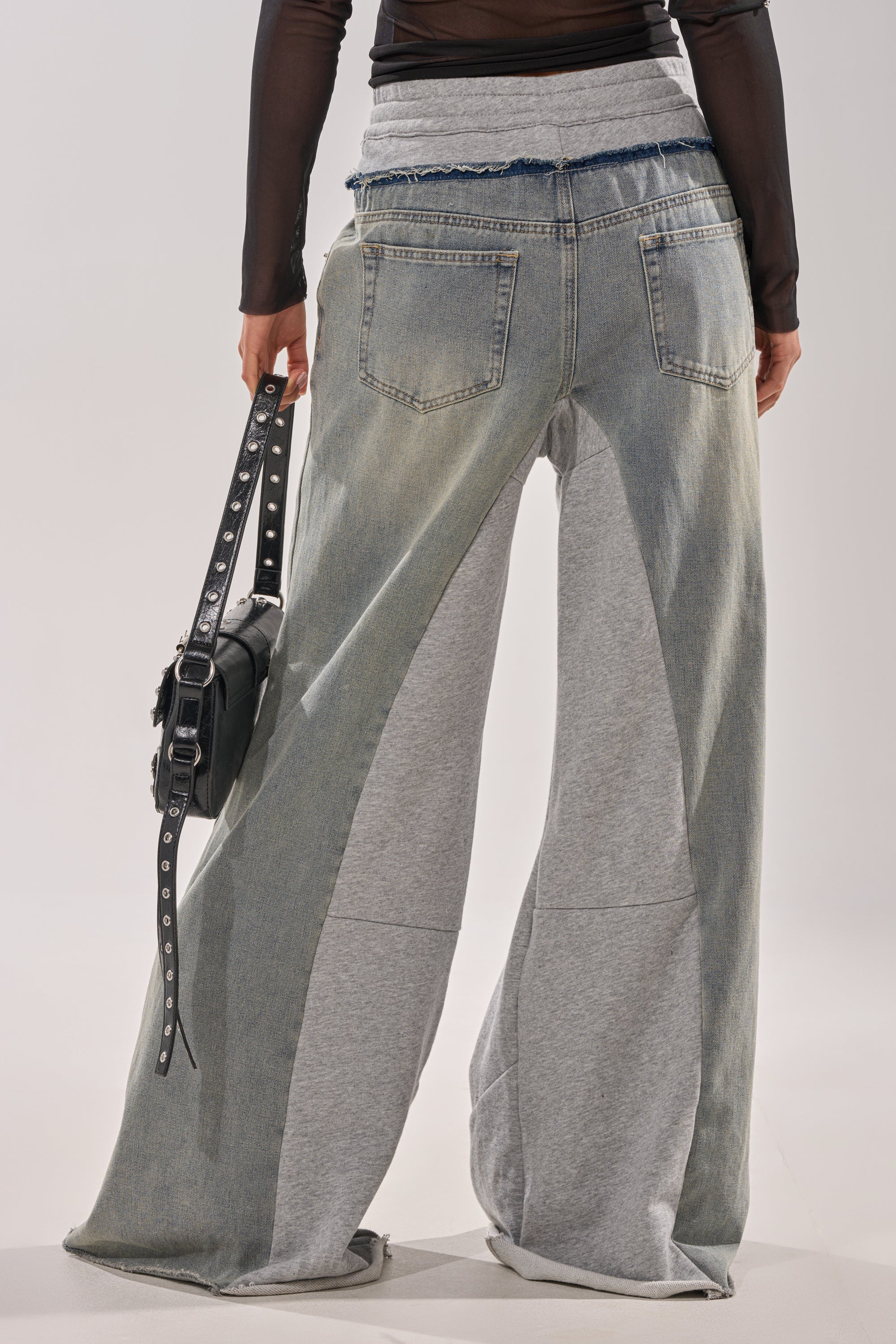 A person faces away, wearing the COMFY WIDE LEG DENIM JOGGER—a hybrid of denim and gray sweatshirt fabric with flared legs—paired with a sheer black long-sleeve top and a black studded bag for an effortless street-style vibe. 4