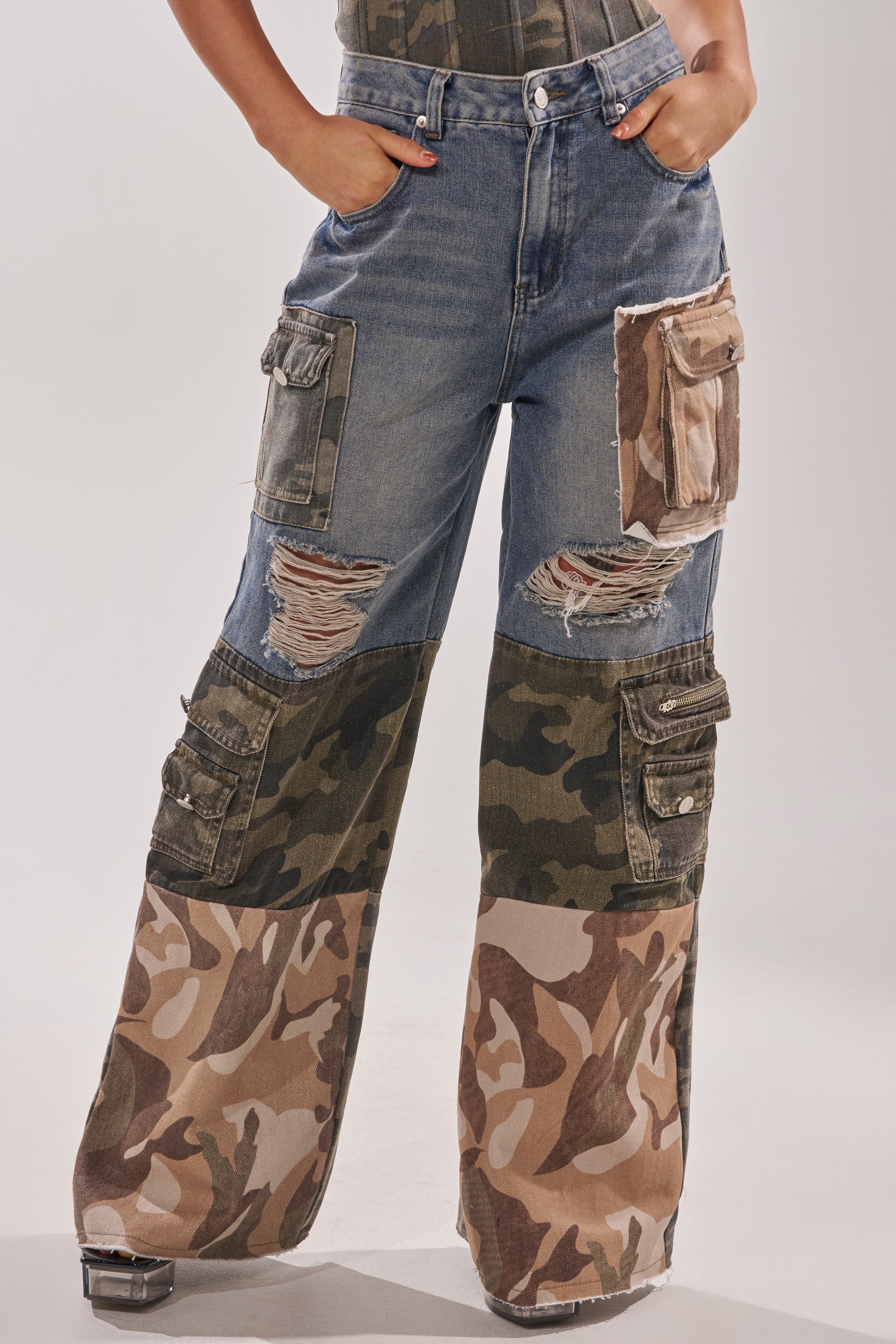 Someone wears the MIXED CAMO DENIM CARGO PANTS—high-waisted, wide-leg streetwear with green and tan camo patchwork, distressed details, and multiple cargo pockets. Their hands rest on their hips. 1