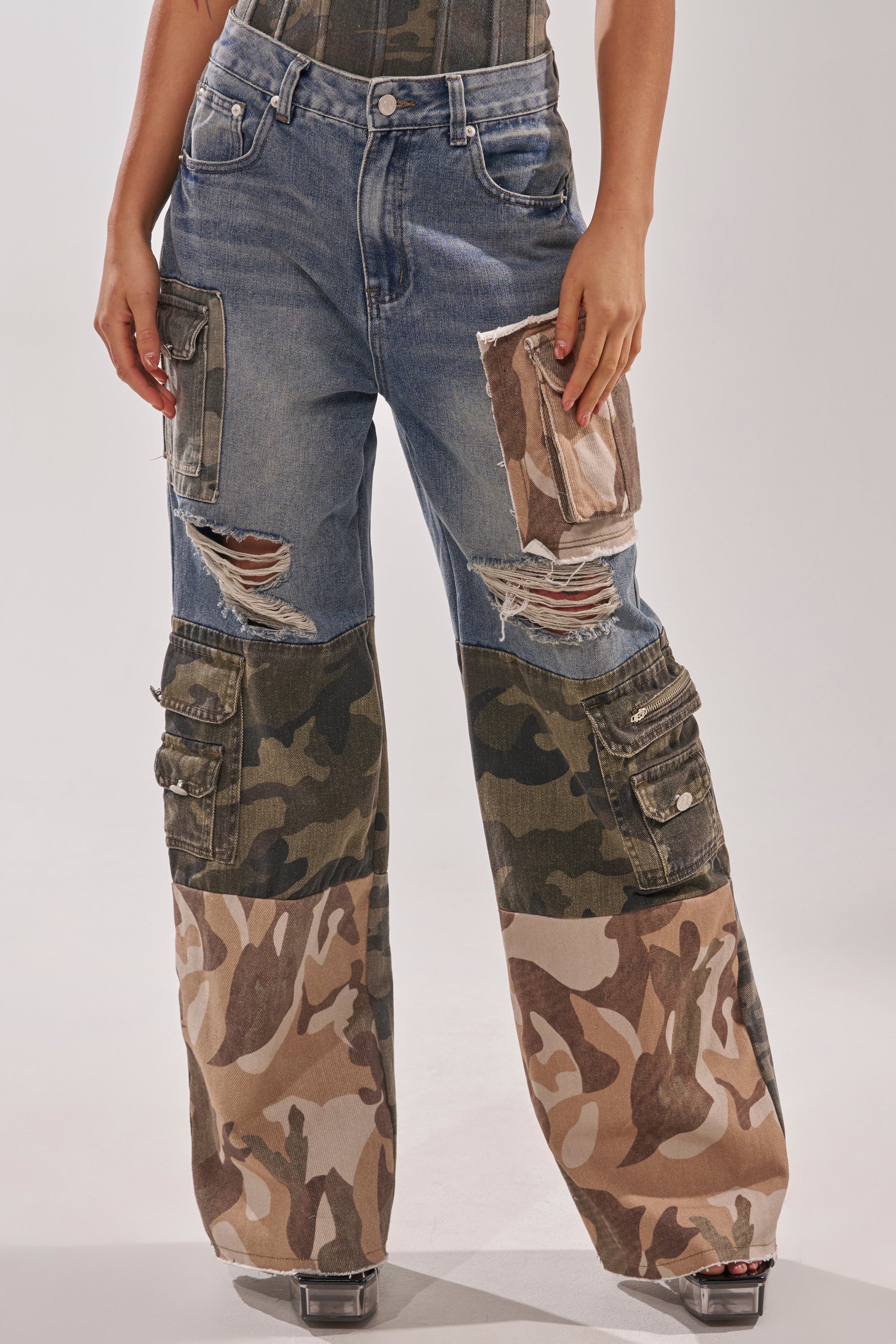 Someone wears the MIXED CAMO DENIM CARGO PANTS, featuring blue denim, distressed details, and camo patchwork panels on the legs, while standing against a plain background. 3