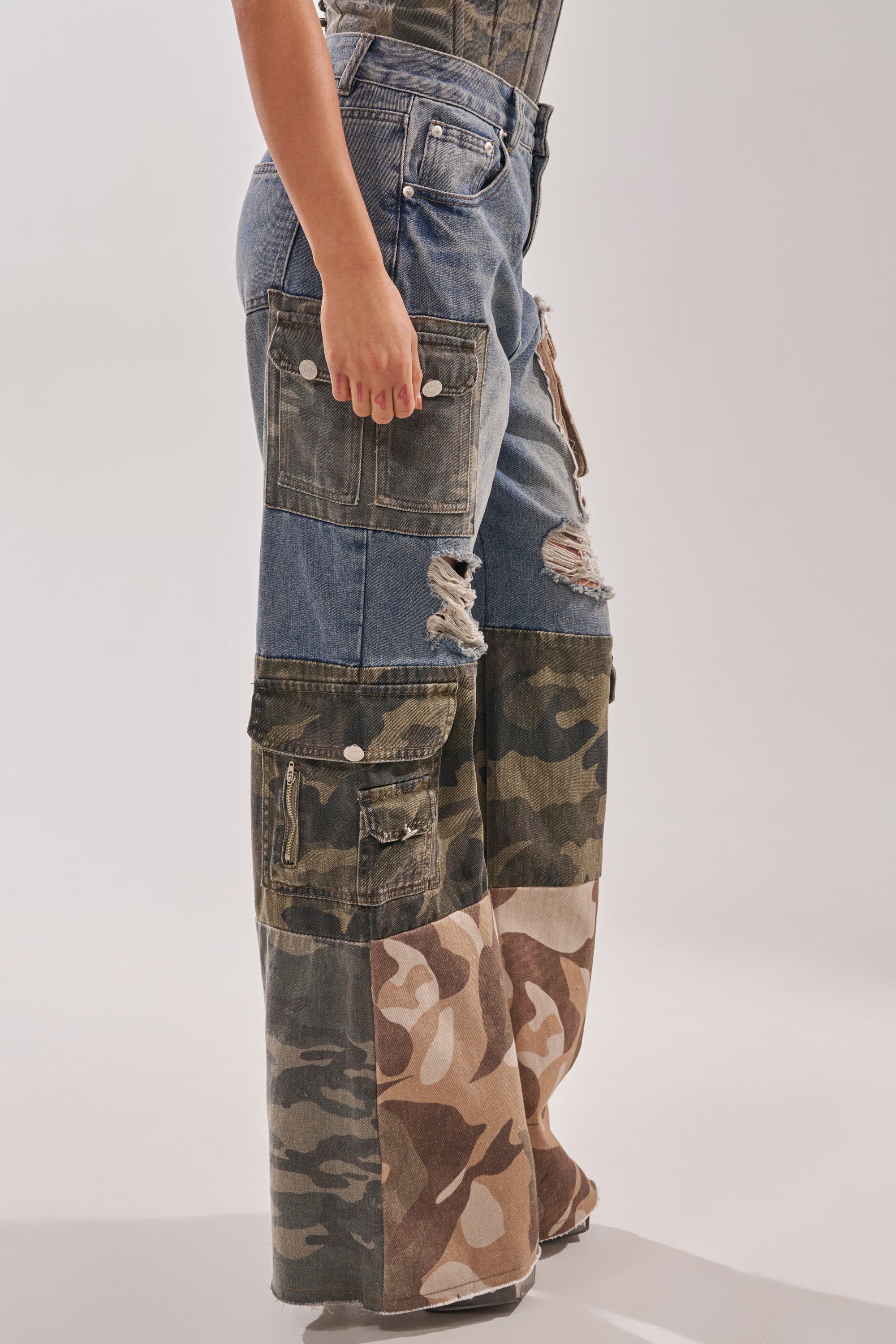 A person wears MIXED CAMO DENIM CARGO PANTS—streetwear-inspired wide-leg cargos made from camo patchwork and distressed denim with large pockets. Only the lower torso and legs appear against a plain background. 4