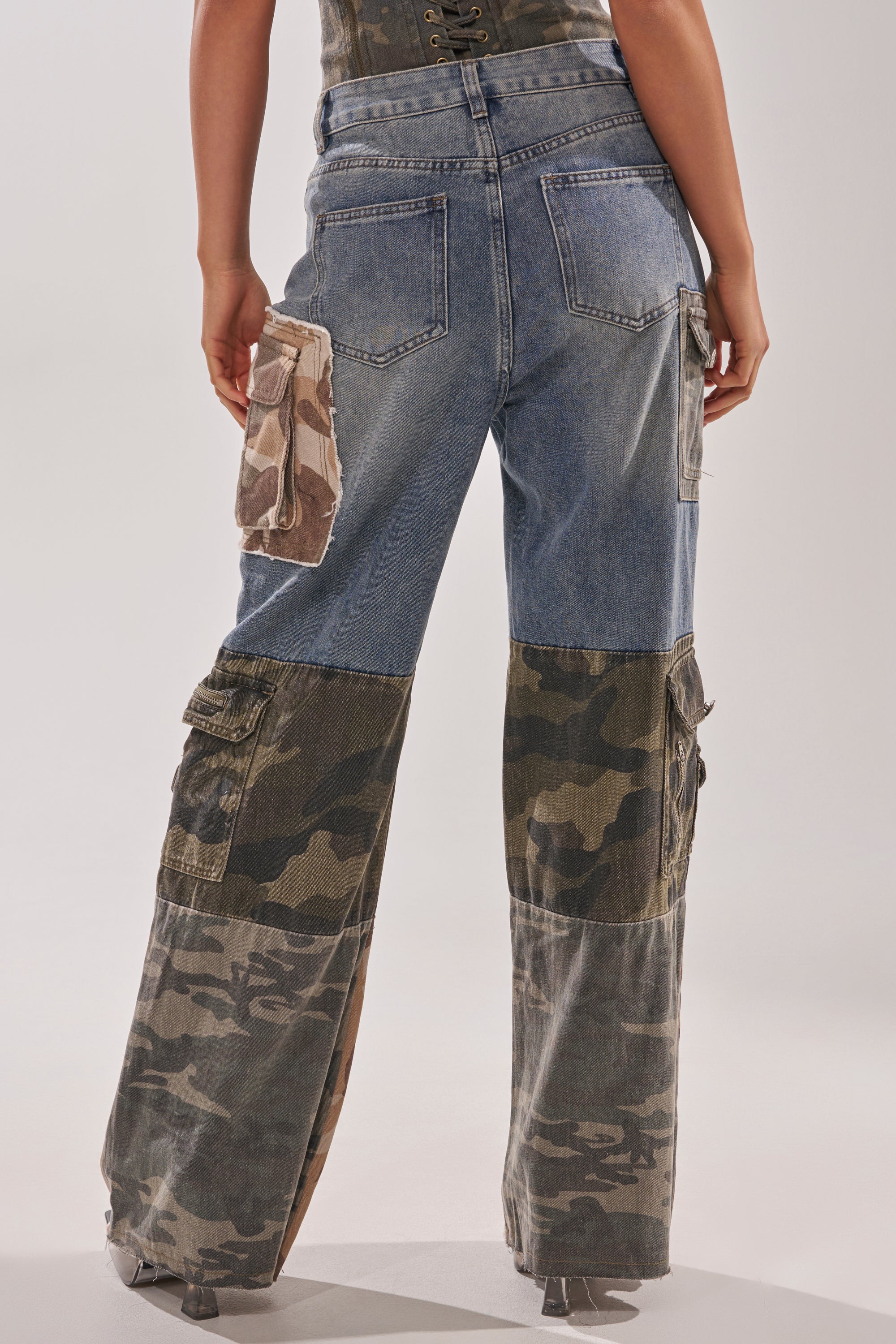 Someone stands facing away, wearing the MIXED CAMO DENIM CARGO PANTS featuring blue denim, camo patchwork panels, and oversized streetwear-style camo pockets on the back and sides. 5