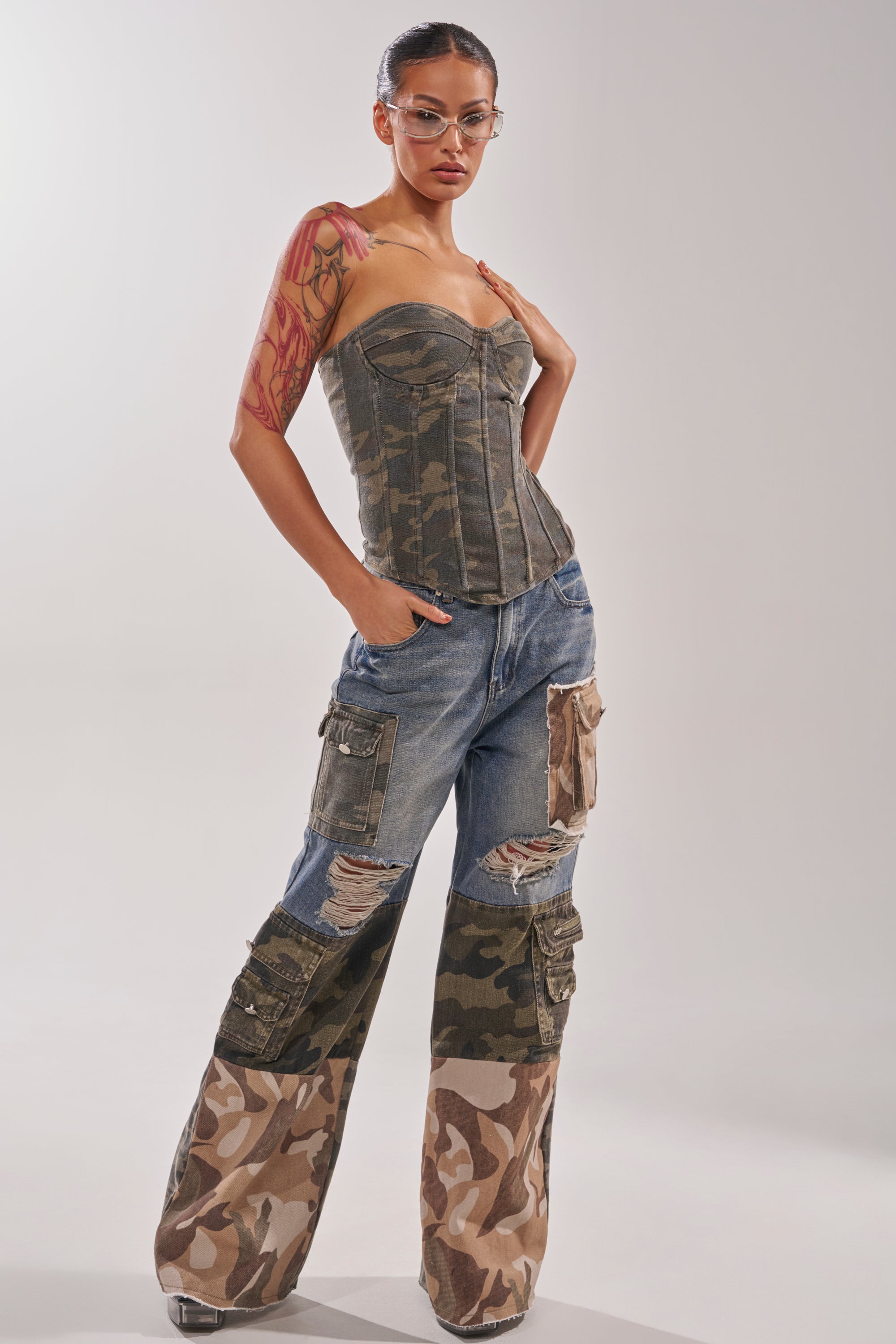 Wearing MIXED CAMO DENIM CARGO PANTS, a camouflage corset, transparent heels, and clear glasses, a person poses confidently against a plain backdrop. 7