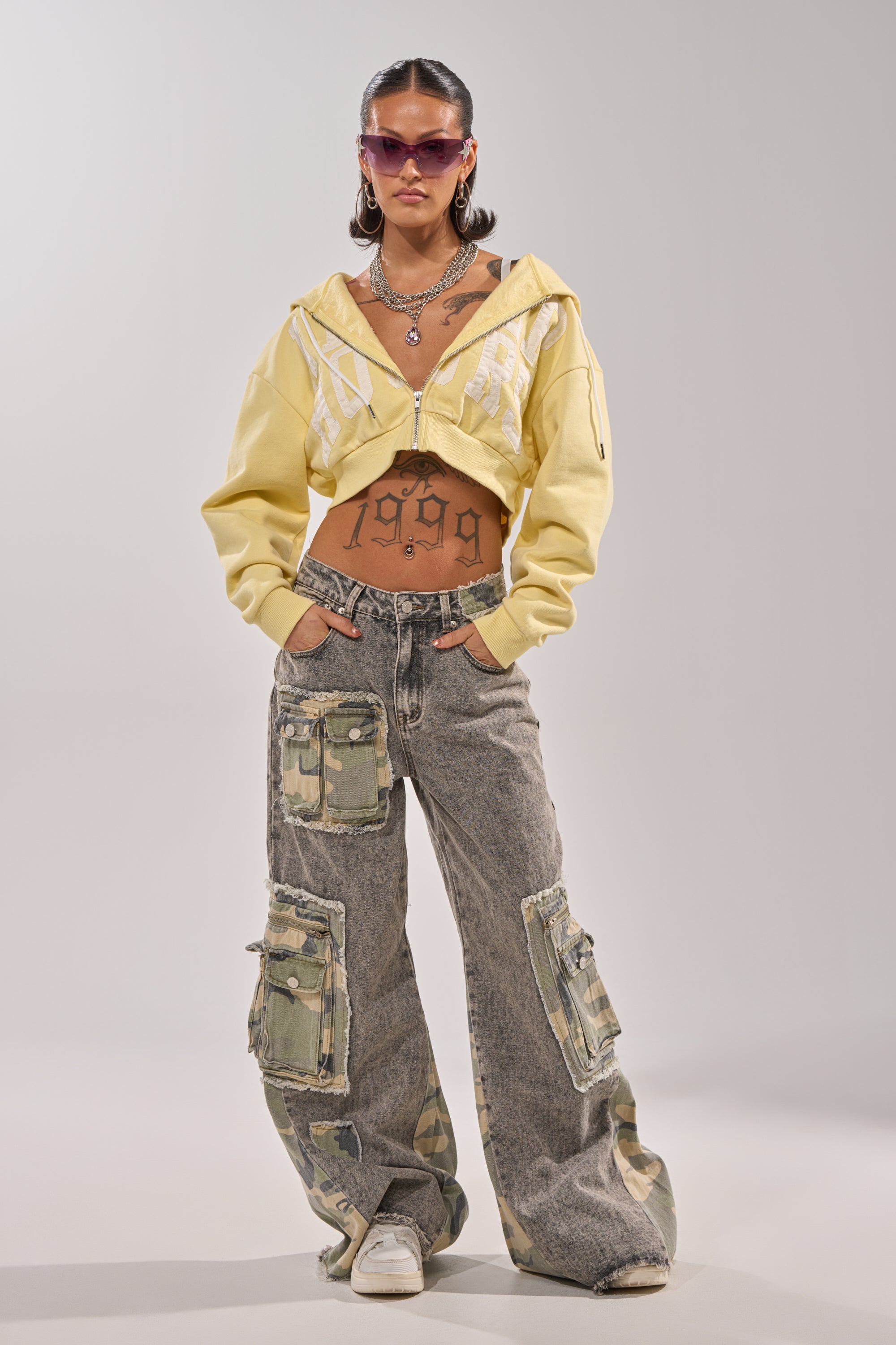 A woman in sunglasses wears the DOWN AND DIRTY CAMO CARGO denim jeans with camo cargo pockets and patches, paired with a yellow cropped hoodie, white sneakers, layered necklaces, and shows off her tattoos against a plain background. 0