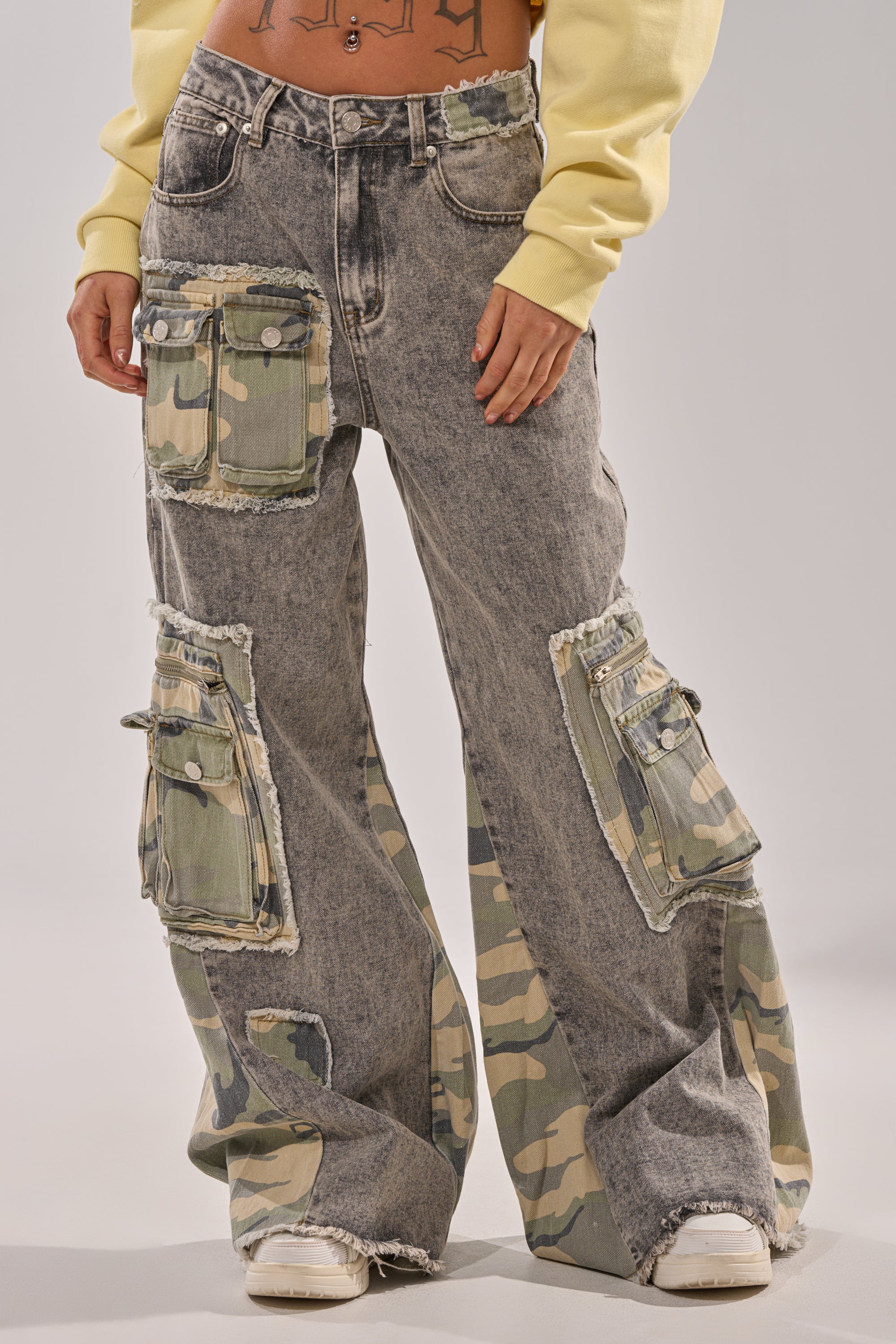 Wearing the DOWN AND DIRTY CAMO CARGO: high-rise waist, acid-wash flared jeans with large camo patch pockets and frayed edges, styled with a yellow cropped sweatshirt and white sneakers. 1