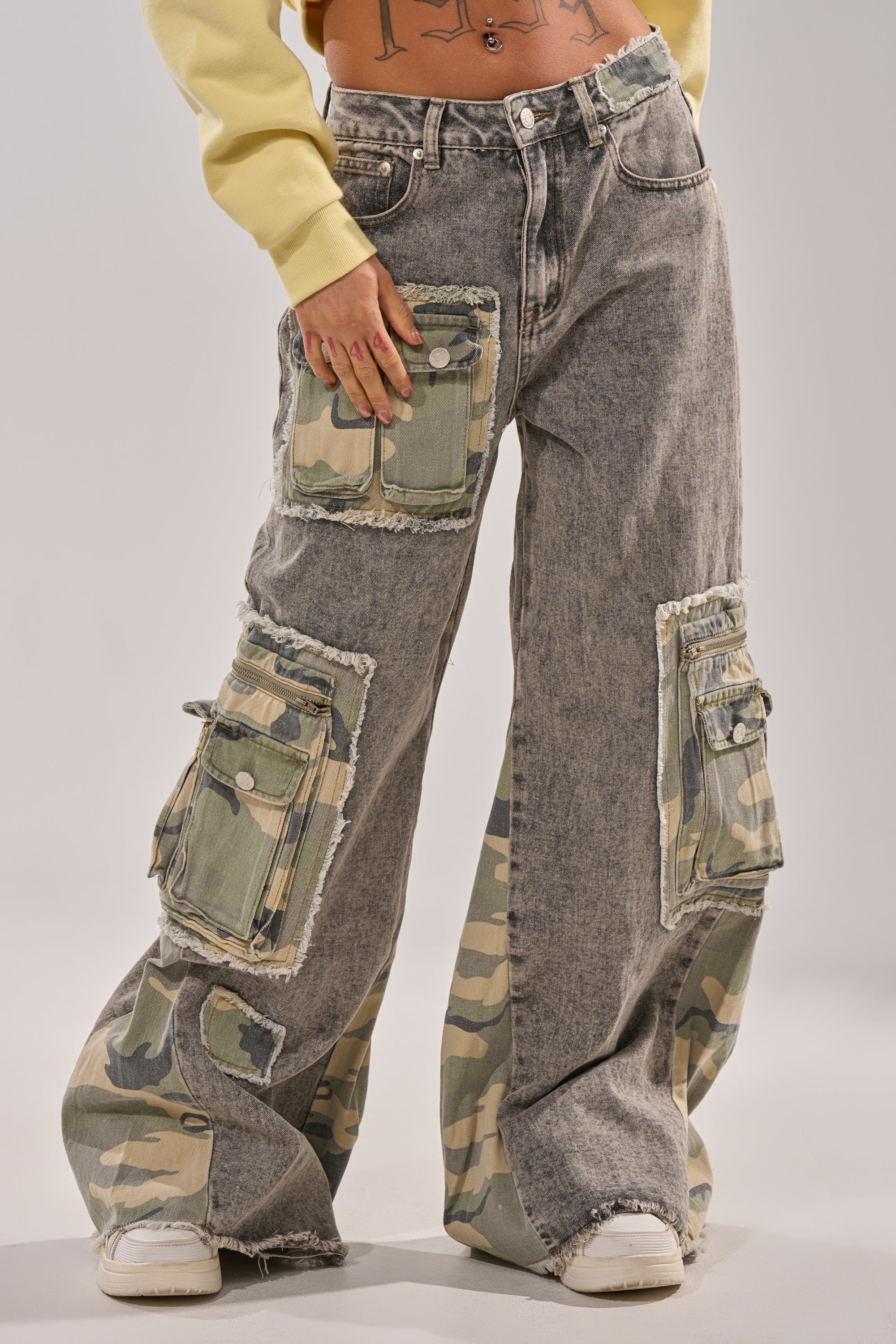 Wearing the DOWN AND DIRTY CAMO CARGO jeans with a high-rise waist and large green camo patch pockets, paired with white sneakers and a yellow top. Torso and head are not visible. 2